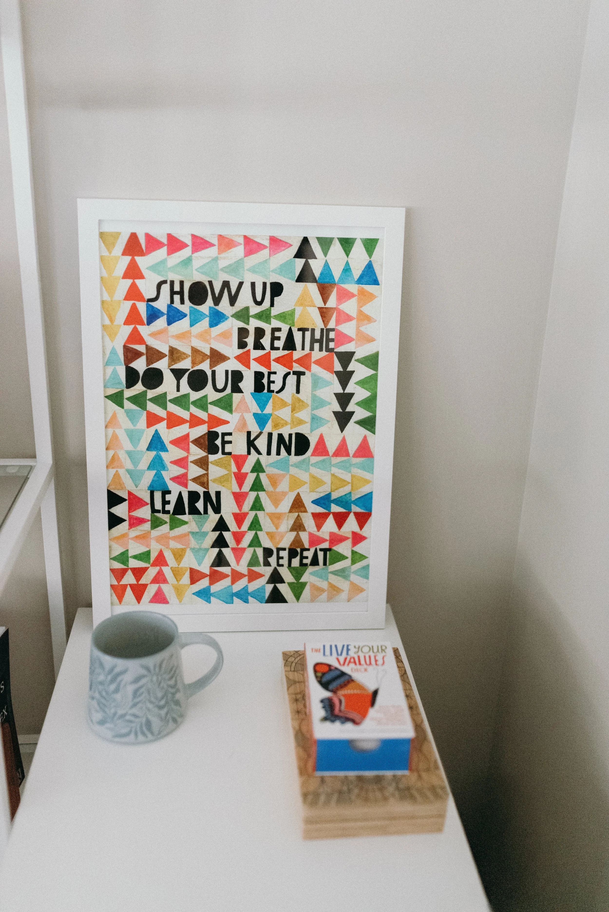 An artsy framed poster with a multitude of colors including red, orange, yellow, green, blue and it reads: Show up, breathe, do your best, be kind, learn and repeat. The poster sits on top of a white cabinet and a floral coffee mug sits in front.
