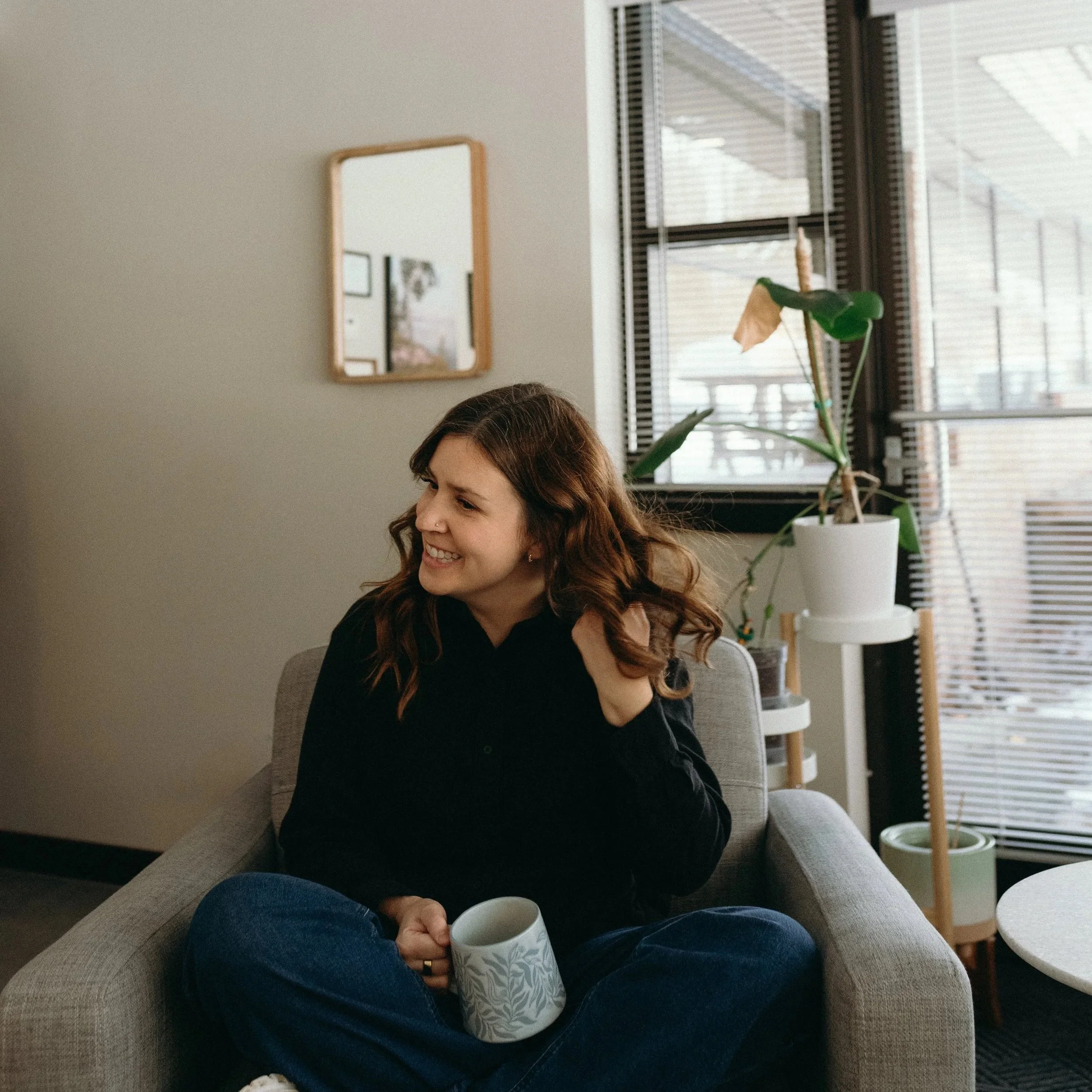 Woman with long brown hair sits cross-legged in a grey chair. She is wearing a black shirt while holding a floral coffee mug.