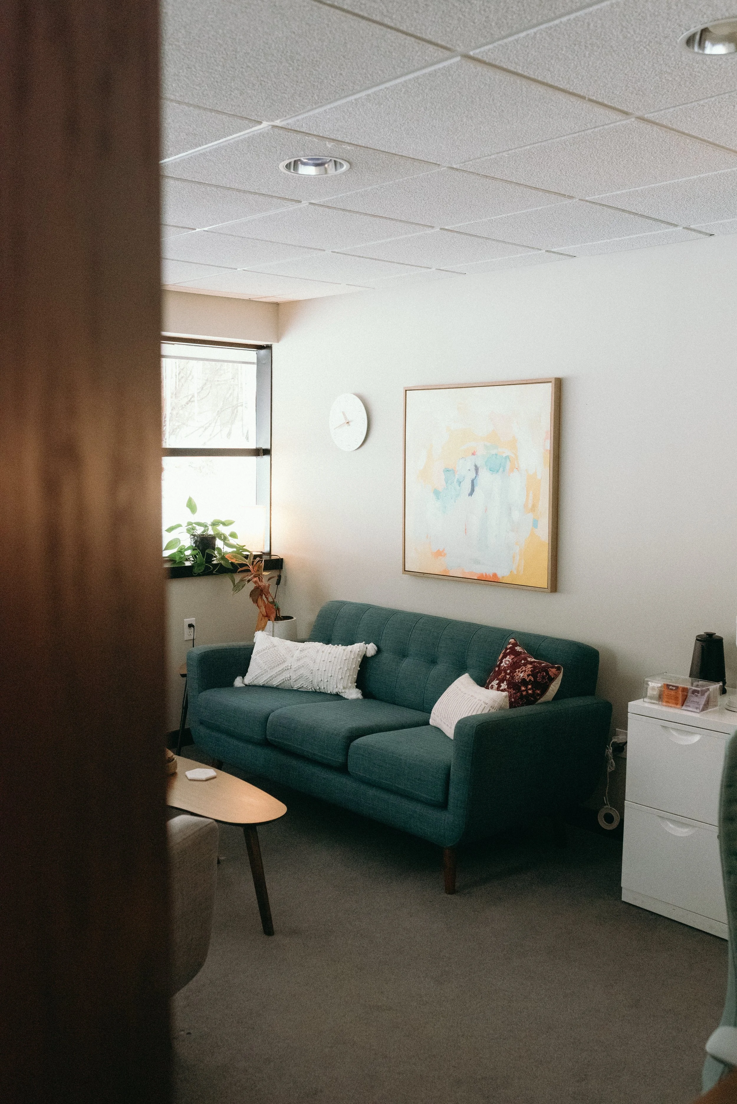 Image of an inviting office with a free couch and white throw pillows. A word coffee table is visible with a green plant in the windowsill along with a soft colored with yellows and blues abstract painting sits on the wall.
