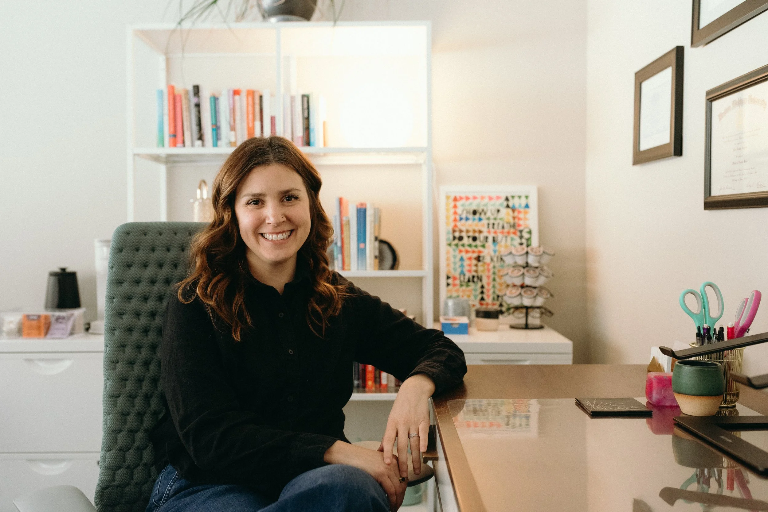 A woman with brown hair sits at her office desk smiling with white bookshelves and an assortment of books behind her.