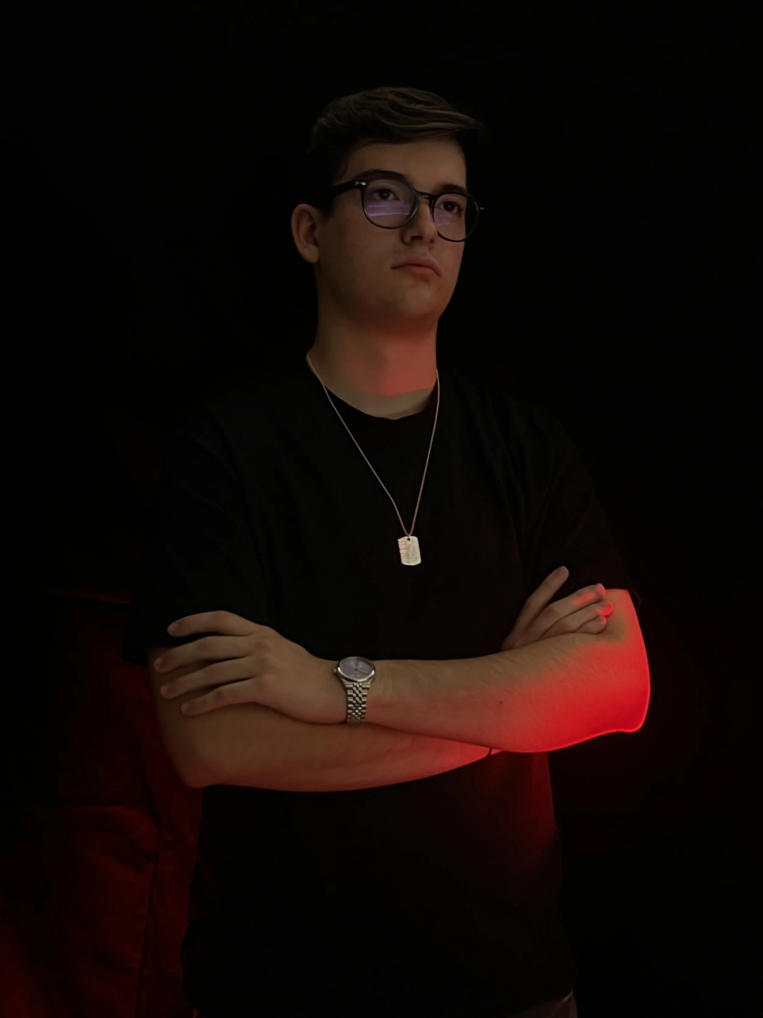 Homem jovem de óculos escuros, camiseta preta, usando relógio e colar, com braços cruzados, de frente para a câmera, em fundo escuro. Luz vermelha ilumina seu braço esquerdo.