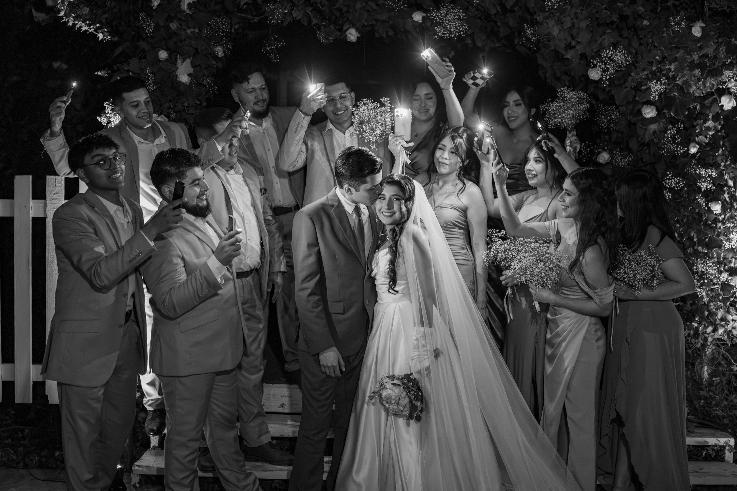 A wedding celebration with the bride and groom kissing, surrounded by friends taking photos at night.