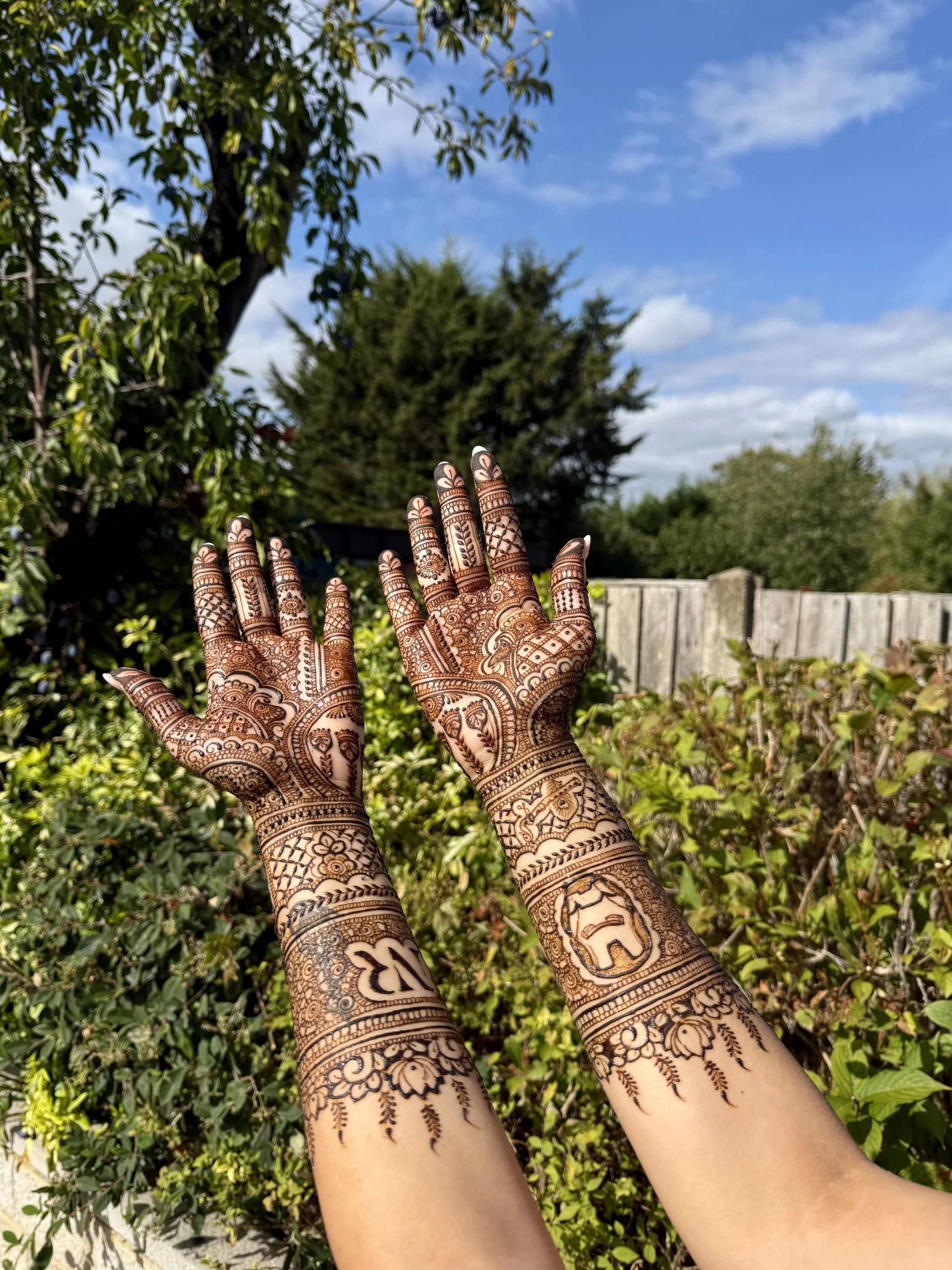 Hands with intricate henna designs raised outdoors against a background of trees, bushes, a wooden fence, and a partly cloudy blue sky.