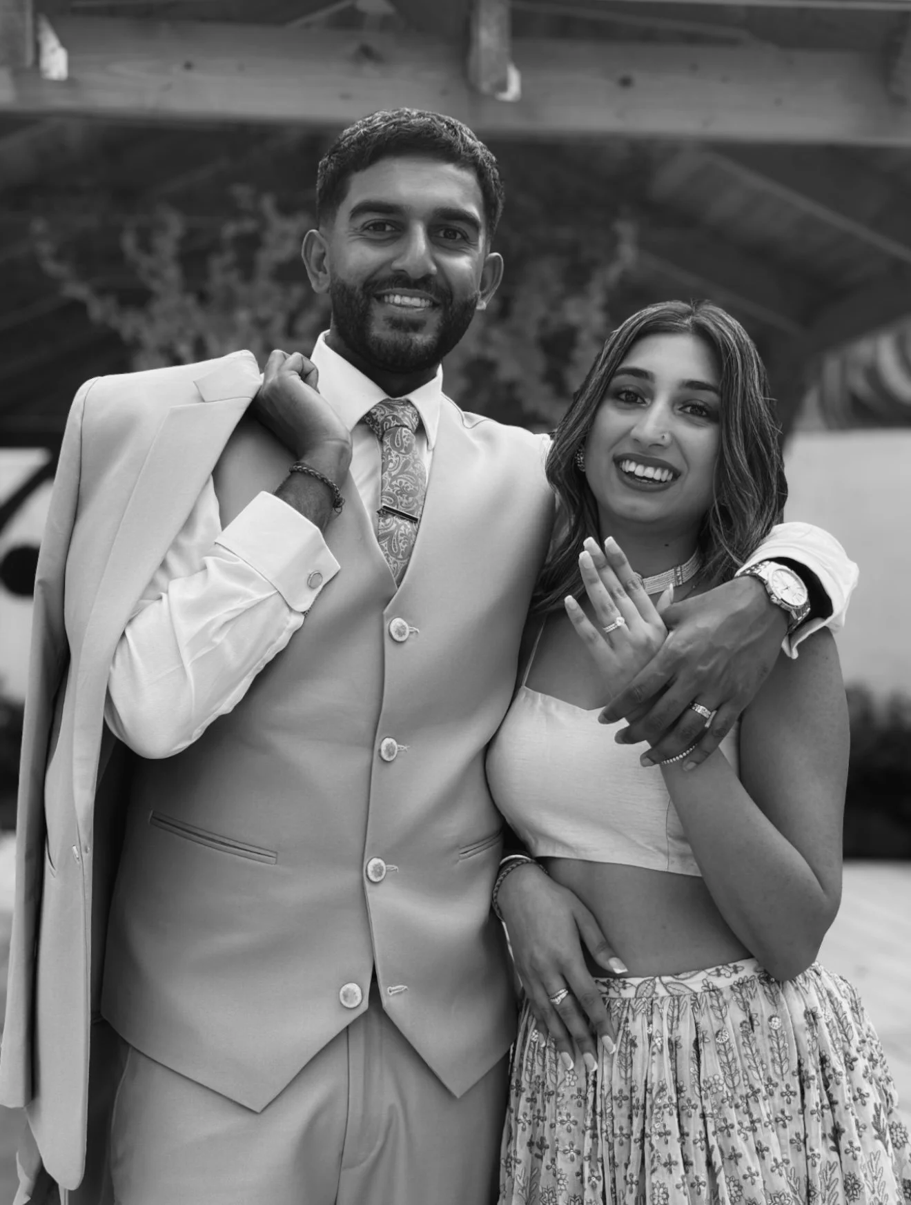 A smiling couple standing close together, the man in a light suit with a patterned tie and the woman in a crop top and patterned skirt, outdoors.