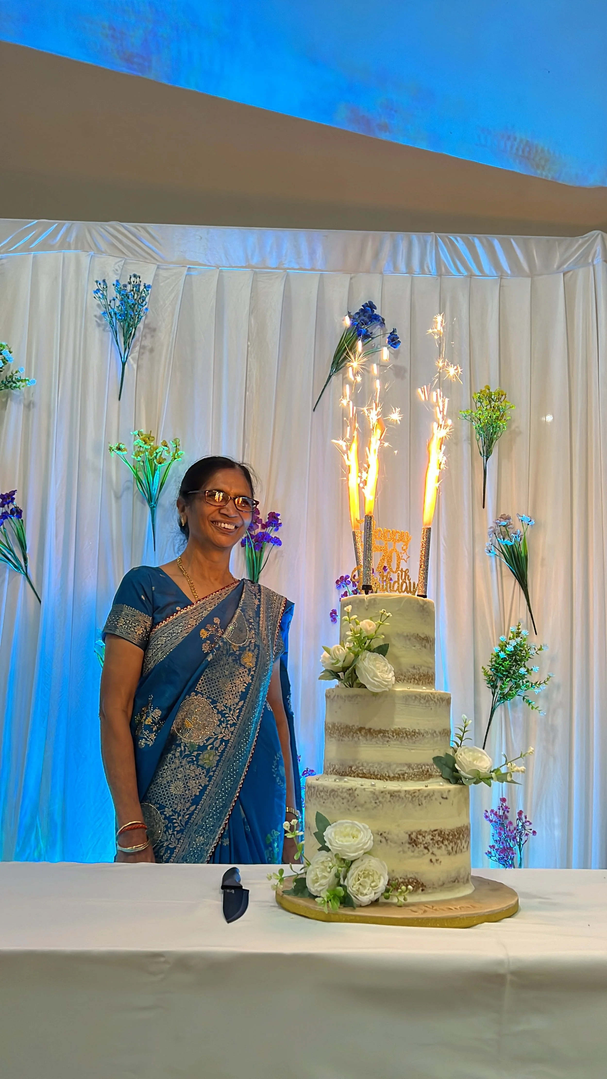 Smiling woman in blue sari standing next to a three-tiered wedding cake decorated with white flowers and sparklers on a decorated table at a celebration.