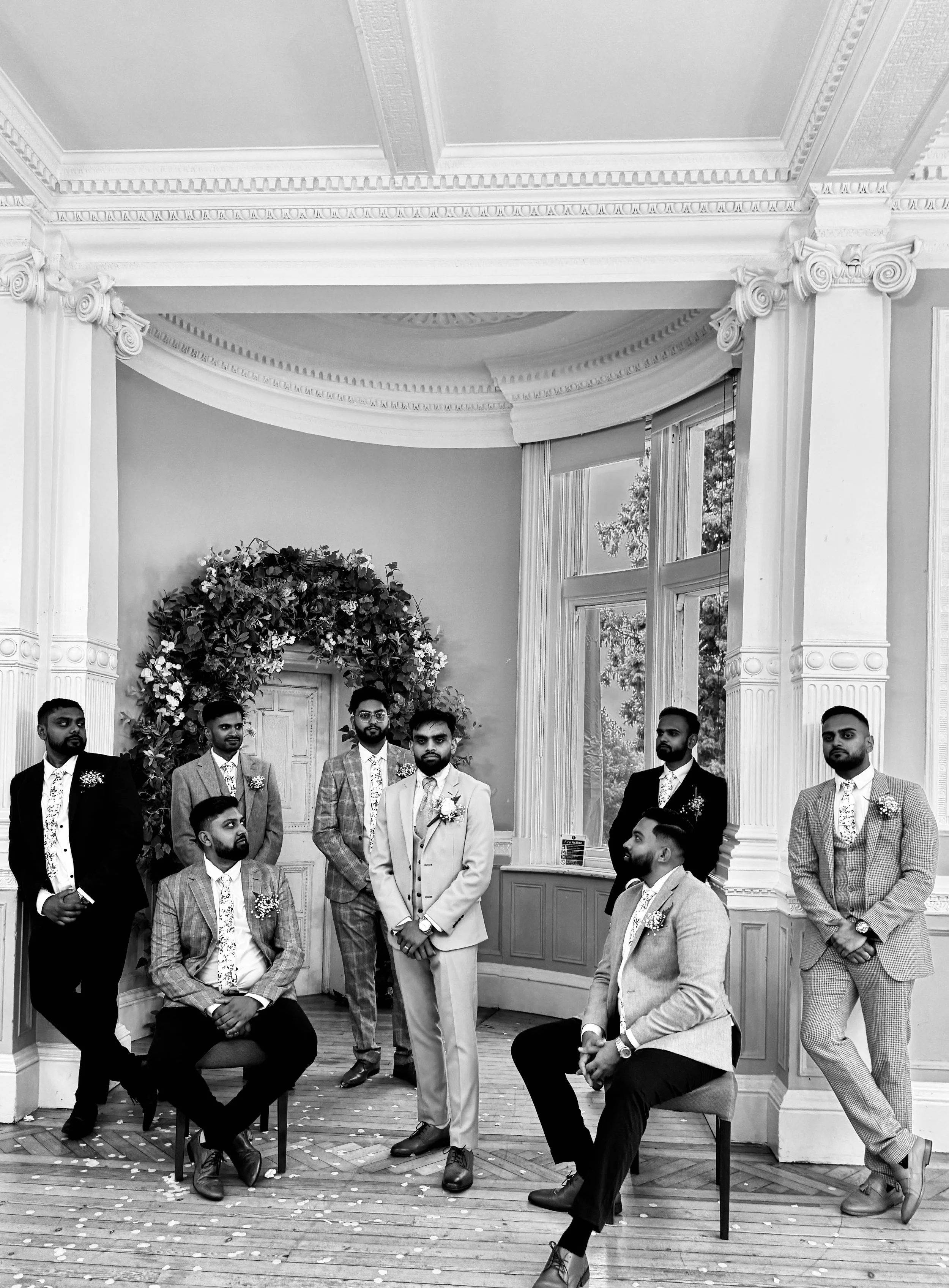 A black and white photo of eight men dressed in suits, some sitting and some standing, inside a room with ornate architectural details and large windows. There is a floral wreath in the background.