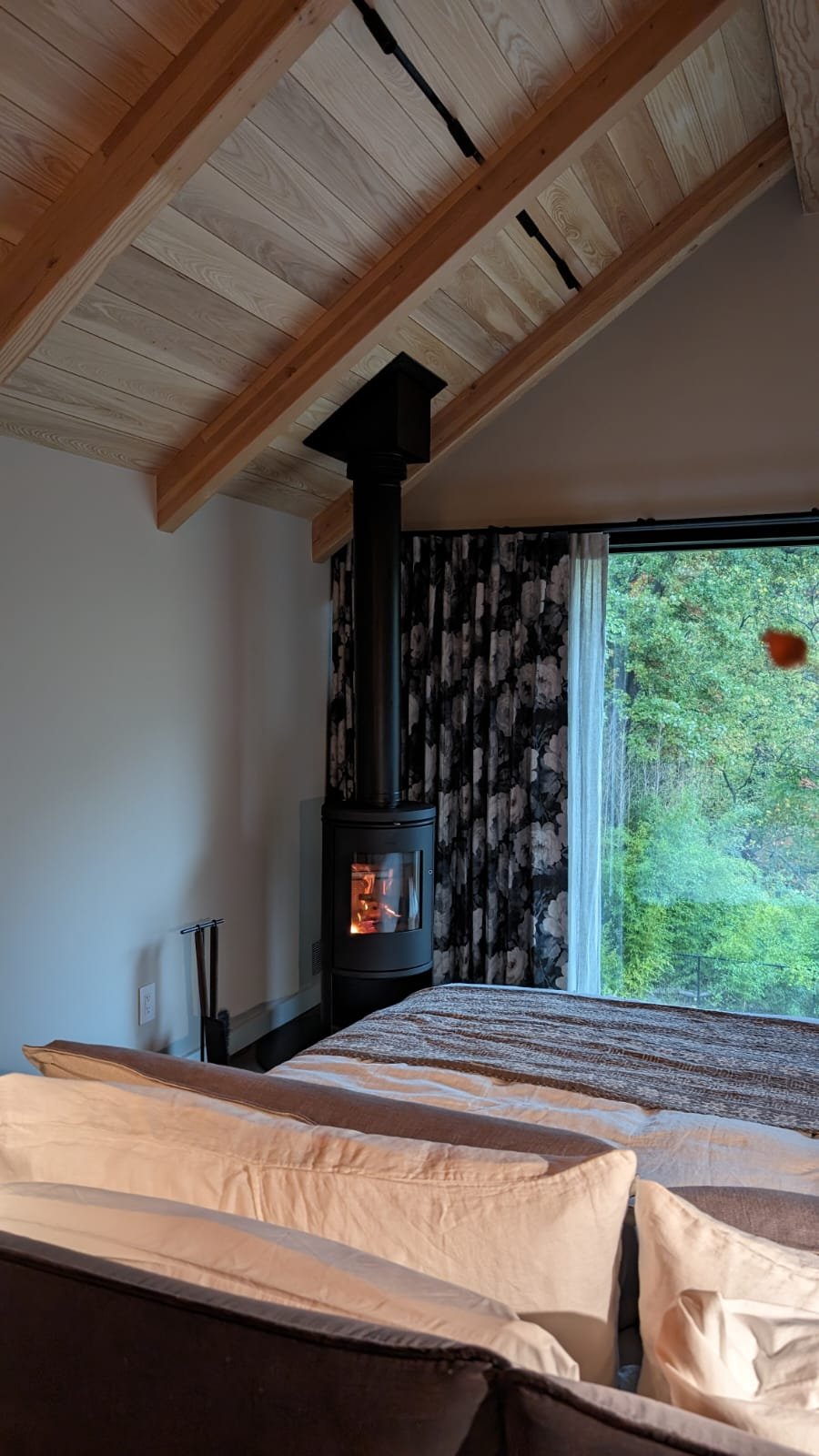 A cozy interior bedroom scene featuring part of a bed with beige pillows, a black wood-burning stove with a visible fire, a large window with dark floral curtains revealing green trees outside, and a wooden ceiling with exposed beams.