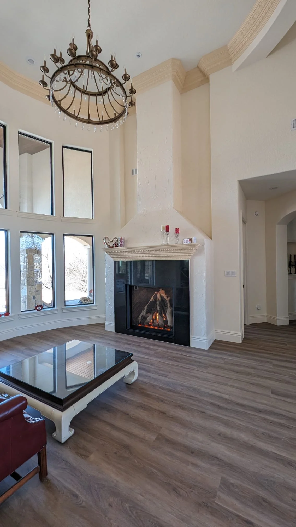 Bright living room with large windows, a tall fireplace with a chimney, and a chandelier hanging from the ceiling. There is a coffee table and a leather armchair in the room.