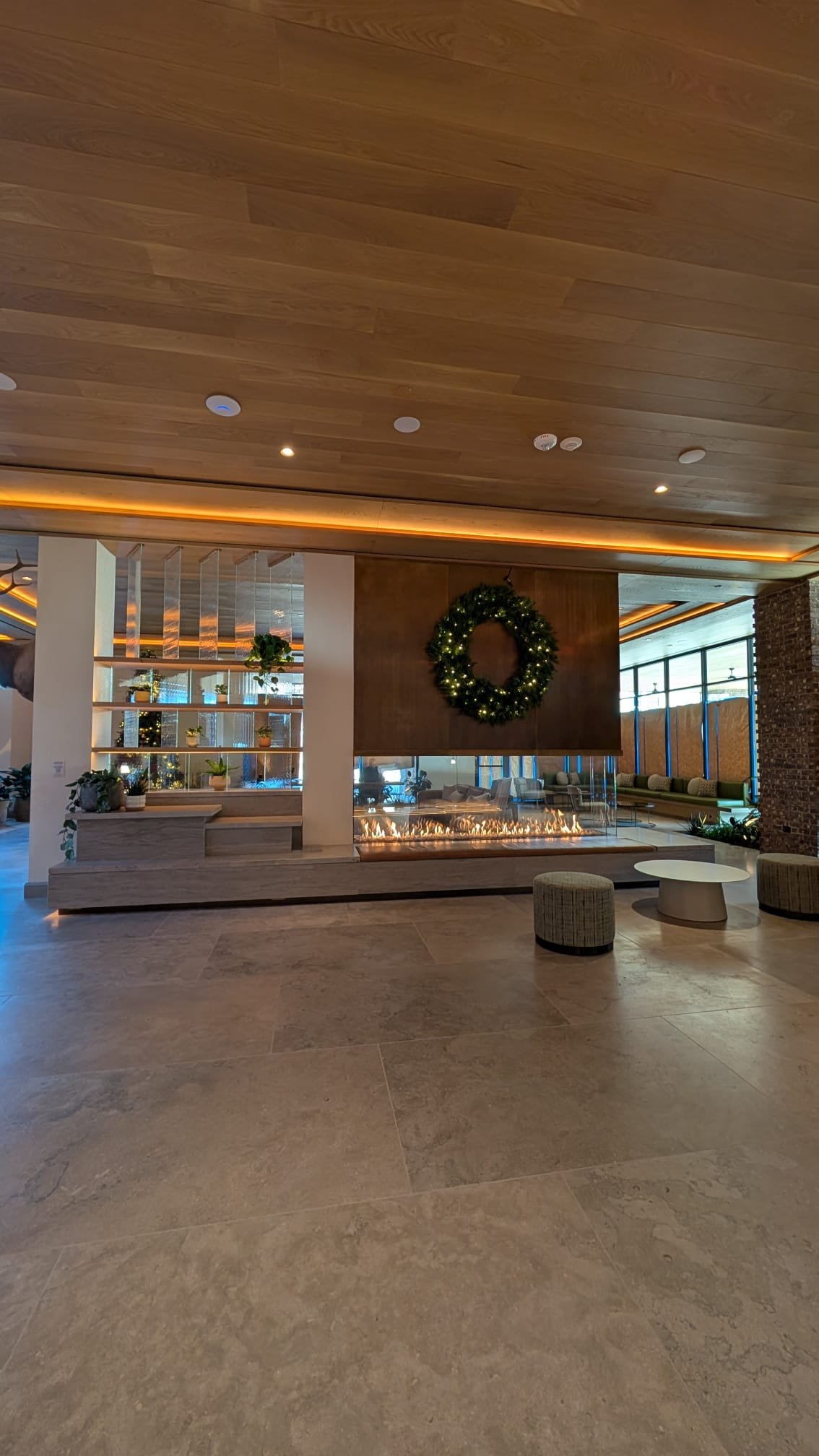A modern hotel lobby with a fireplace decorated for the holidays, featuring a green wreath with lights, and a shelf with potted plants. There are seating areas and large windows in the background.