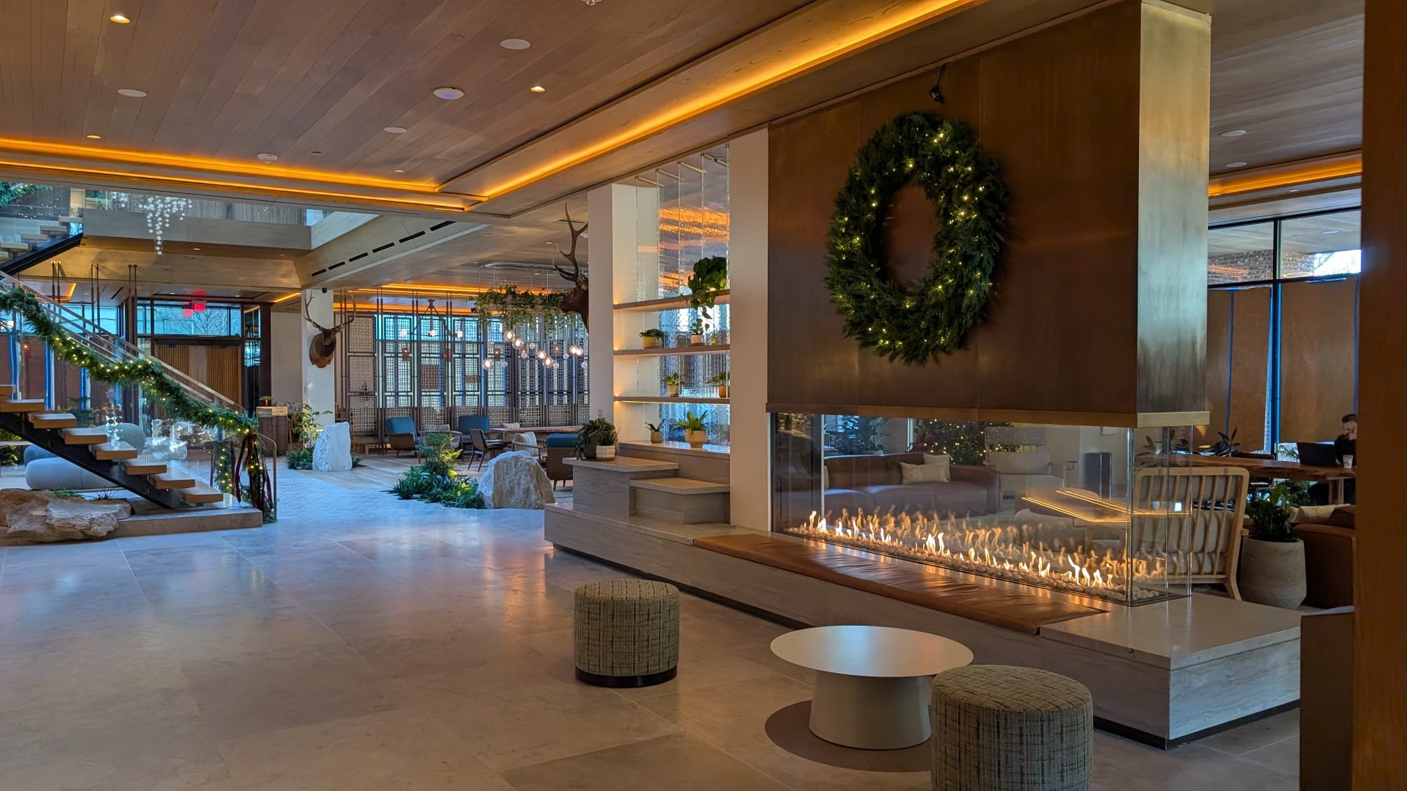 A modern hotel lobby decorated for the Christmas season with a wreath on the wall, a fireplace, and festive garland on the staircase. The space features wooden ceiling and wall accents, large windows, and seating areas with tables and plants.