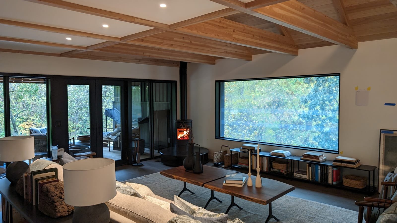 Modern living room with wood-beam ceiling, large window showing trees, coffee table, bookshelves, and a wood stove.