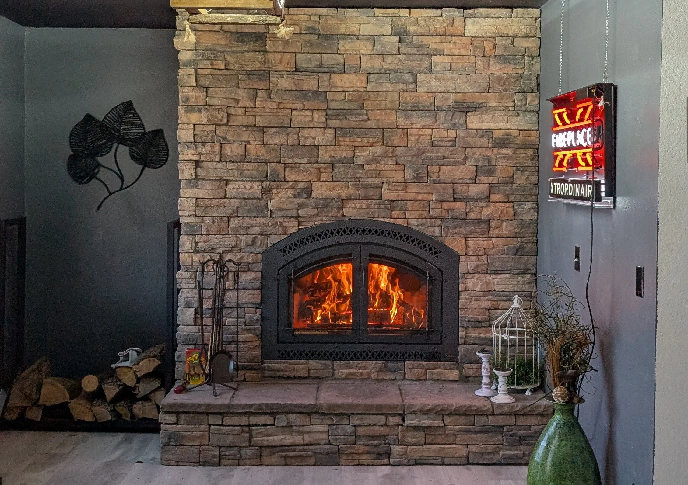 A cozy living room corner featuring a stone fireplace with a burning fire behind a black, arched metal door. To the left, stacked firewood is placed on the floor, and to the right, decorative items including white candles, a wire birdcage, and a green vase with dried plants. An art piece of black leaves is on the dark gray wall to the left, and a neon sign with red and white text is mounted on the wall to the right.