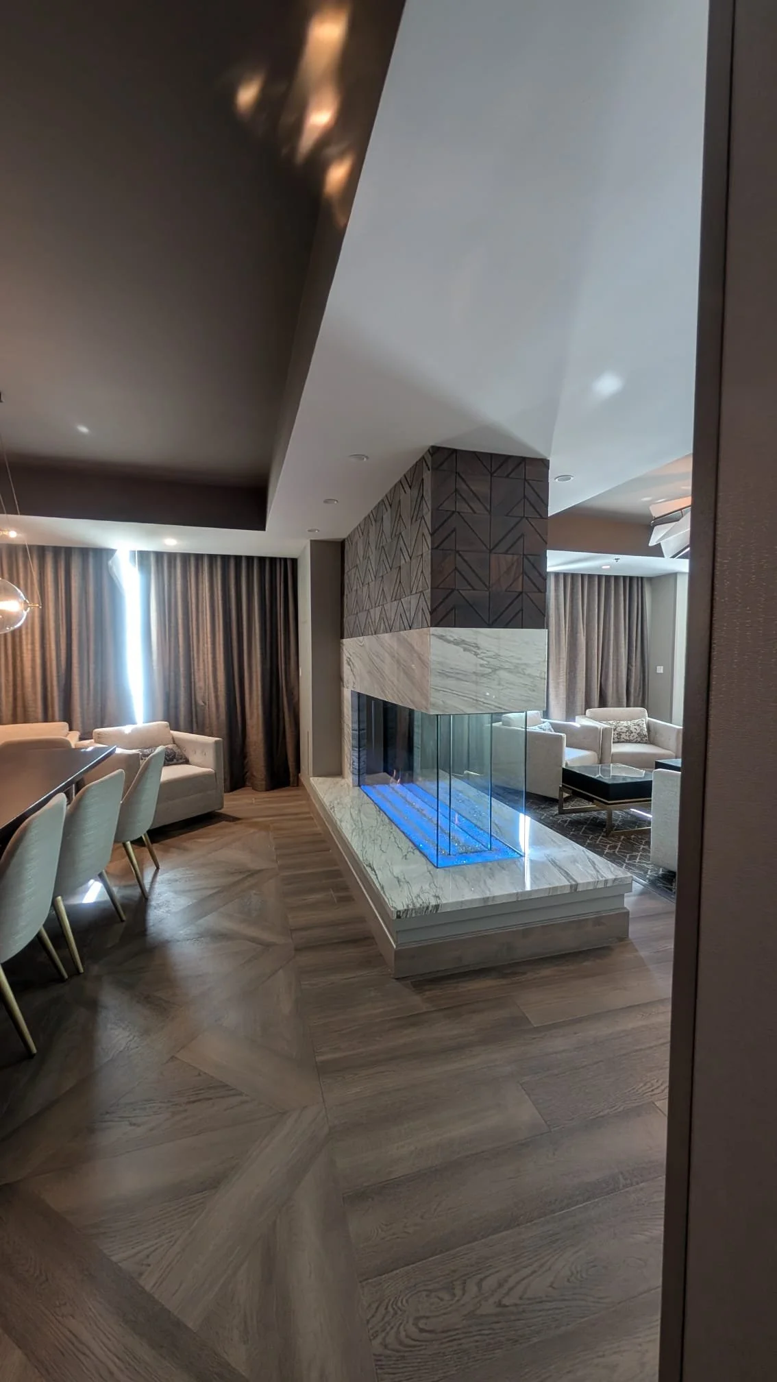 Modern living room with a fireplace, beige sofas, and large curtains, with wooden flooring and ambient lighting.