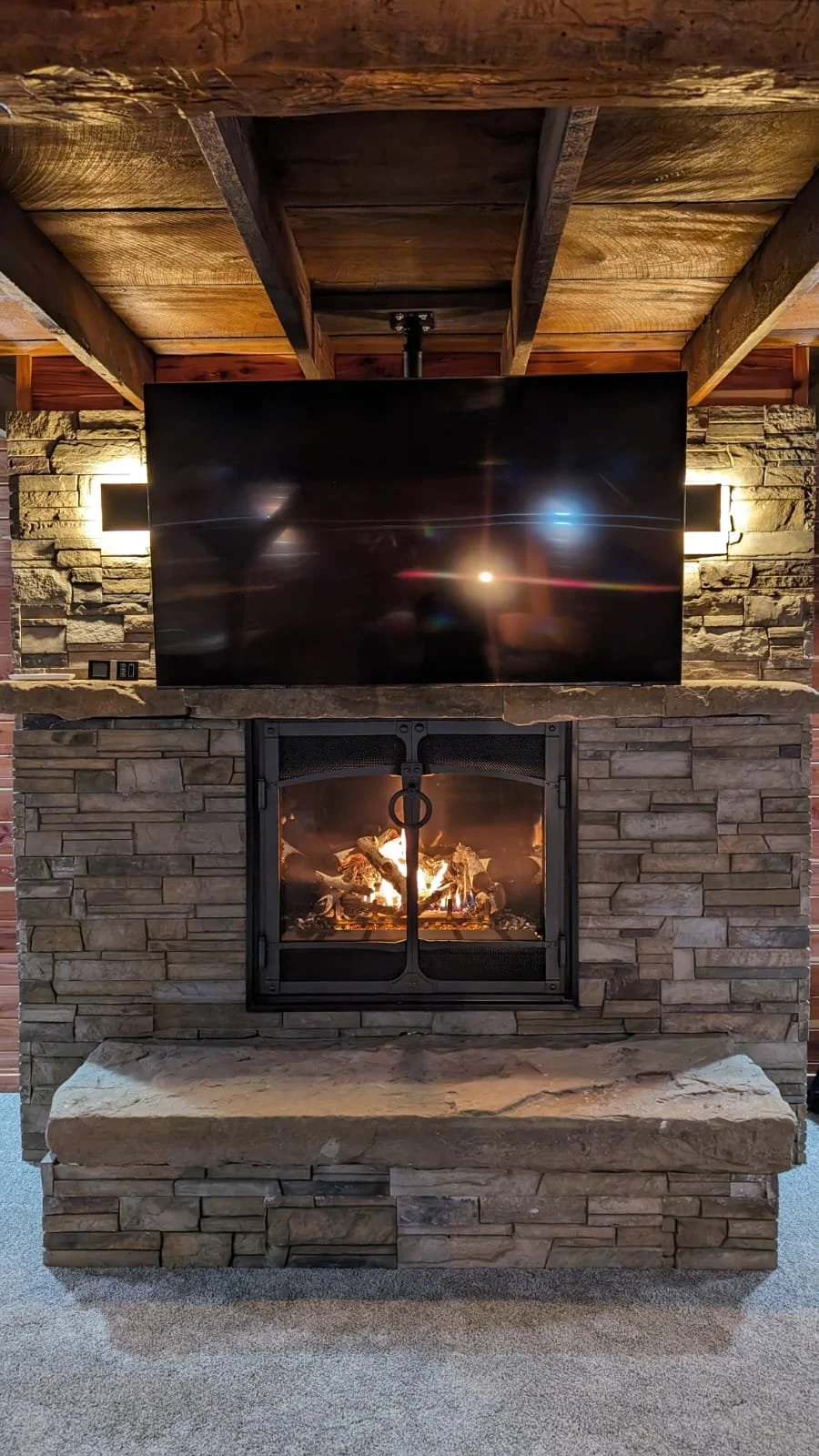 Living room fireplace with a stone surround, a fire burning inside, and a mounted flat-screen TV above it, with wooden beams on the ceiling.