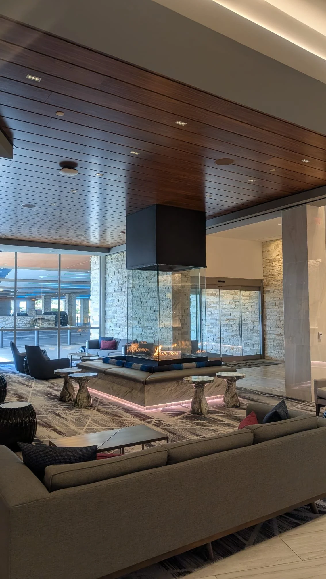 Modern hotel lobby with a central fireplace, seating area with sofas and chairs, large windows, and a wood-paneled ceiling.