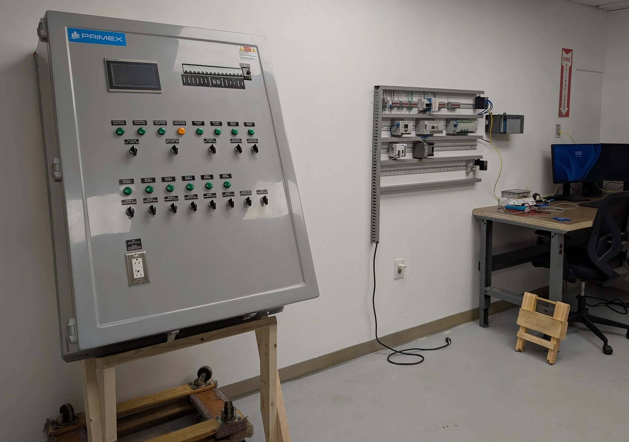 Electrical control panel with switches and buttons in an industrial or laboratory setting, nearby a work desk with electronic equipment and a computer monitor.