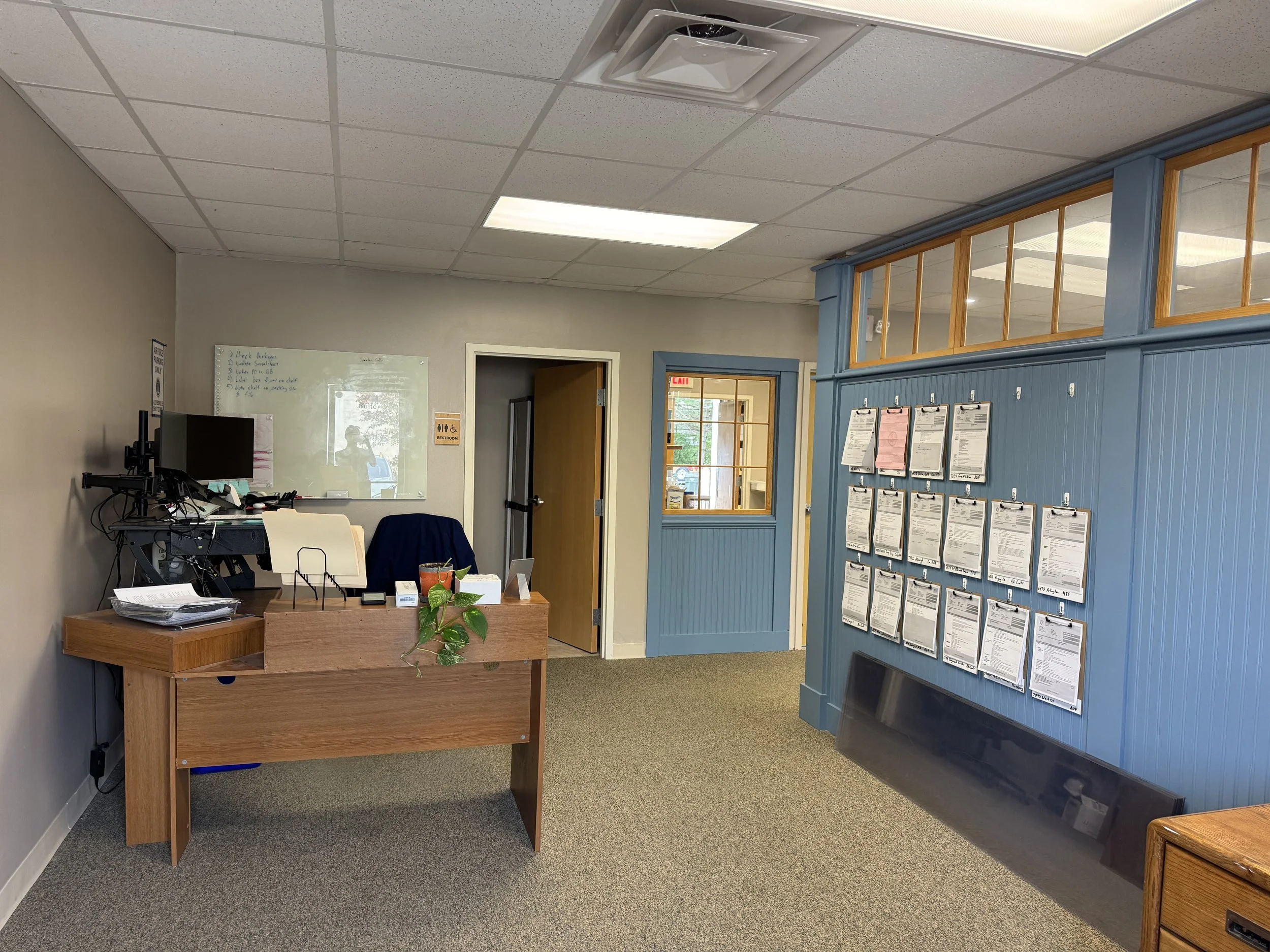 Office reception area with a wooden desk, computer, and papers, blue wall with clipboards displaying documents, door with glass window, and a whiteboard with writing on it.