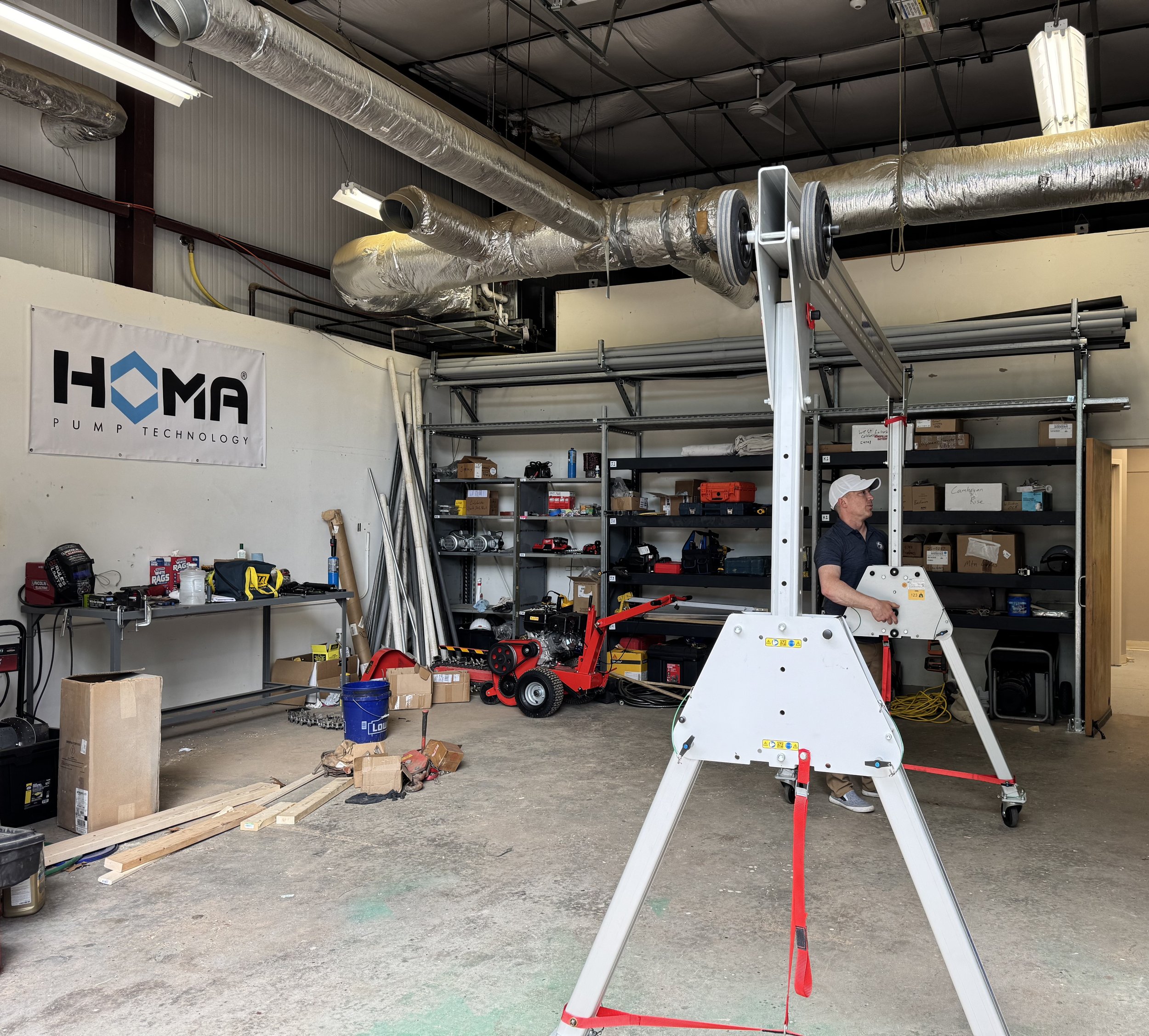 A workshop space with tools and equipment, a person operating a piece of construction machinery, shelves with boxes and supplies, a large sign that reads 'HOMA Pump Technology', and exposed ductwork on the ceiling.