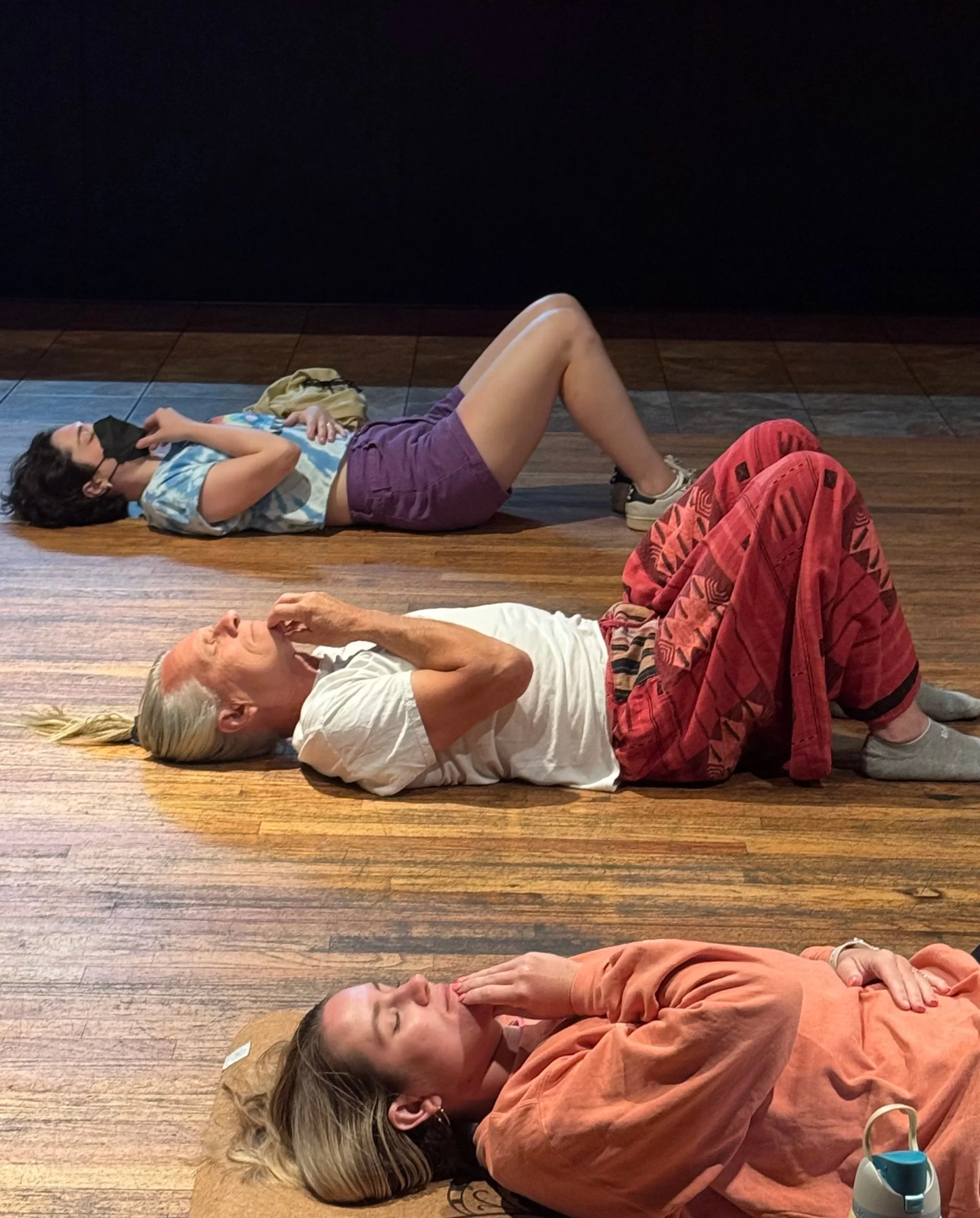 Three people in a relaxed somatic movement pose on a wooden floor