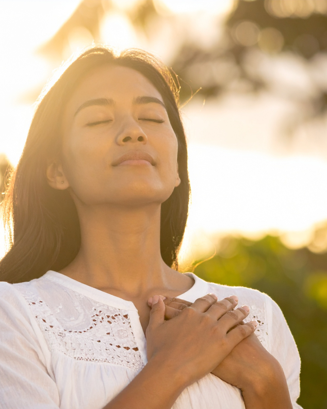 A woman with closed eyes and a peaceful expression, holding her hands over her heart, outdoors with sunlight in the background.