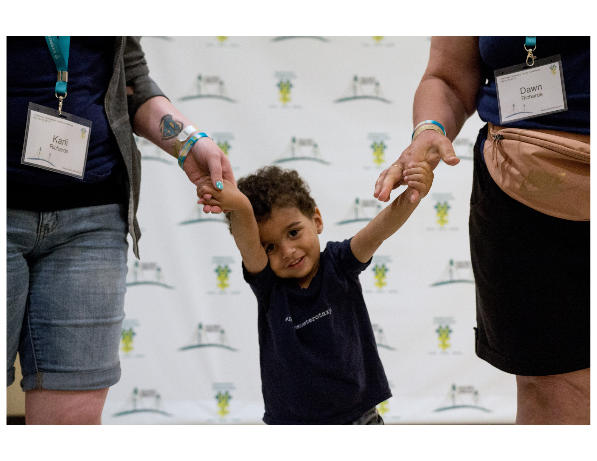 A smiling toddler with light brown skin and dark, curly hair stands between two adults, holding one of their hands with each of his. The adults are only partially visible with the focus being on the little cutie.