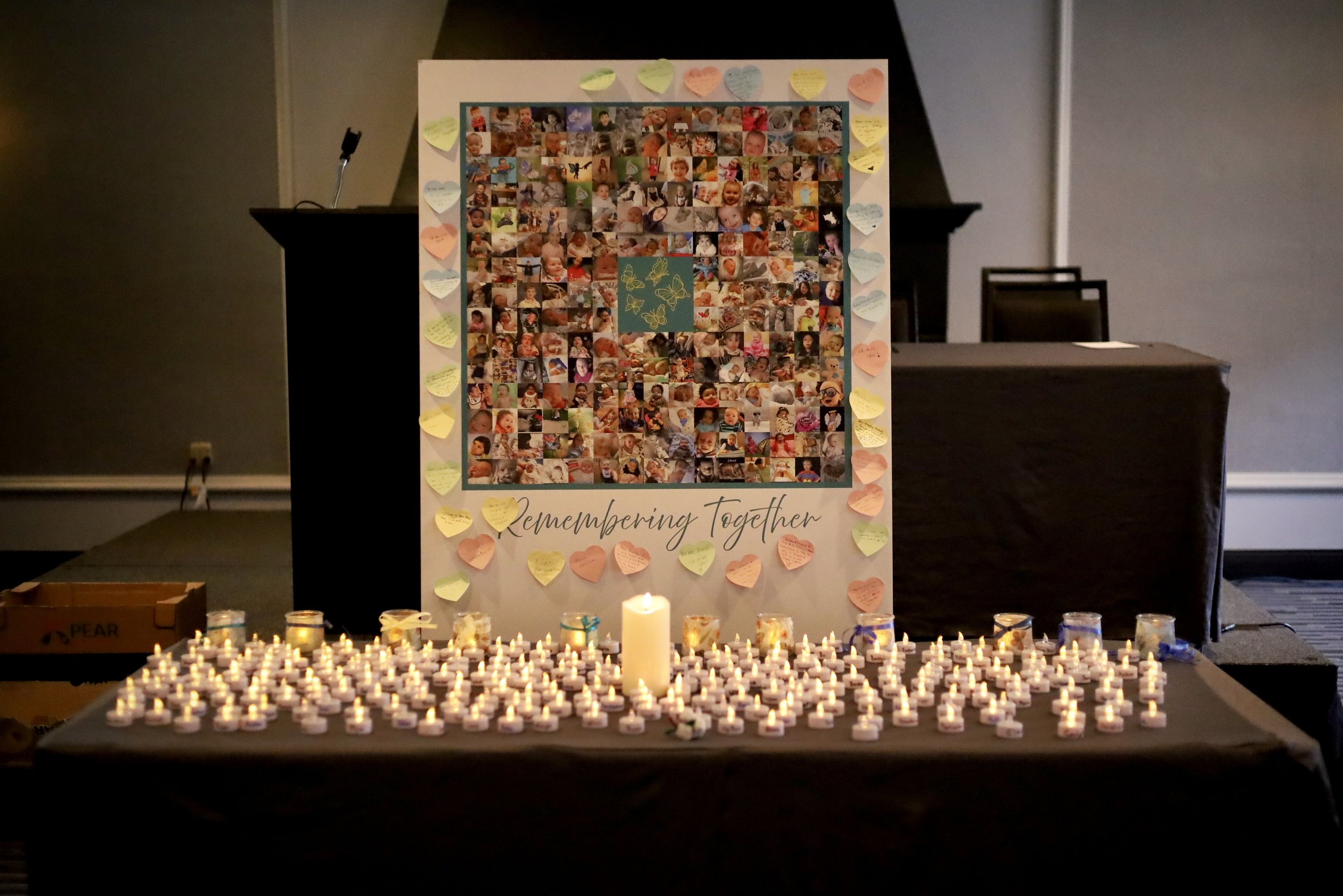 a large collage of faces with the text "remembering together" stands on a table covered in lit tea lights and one large candle