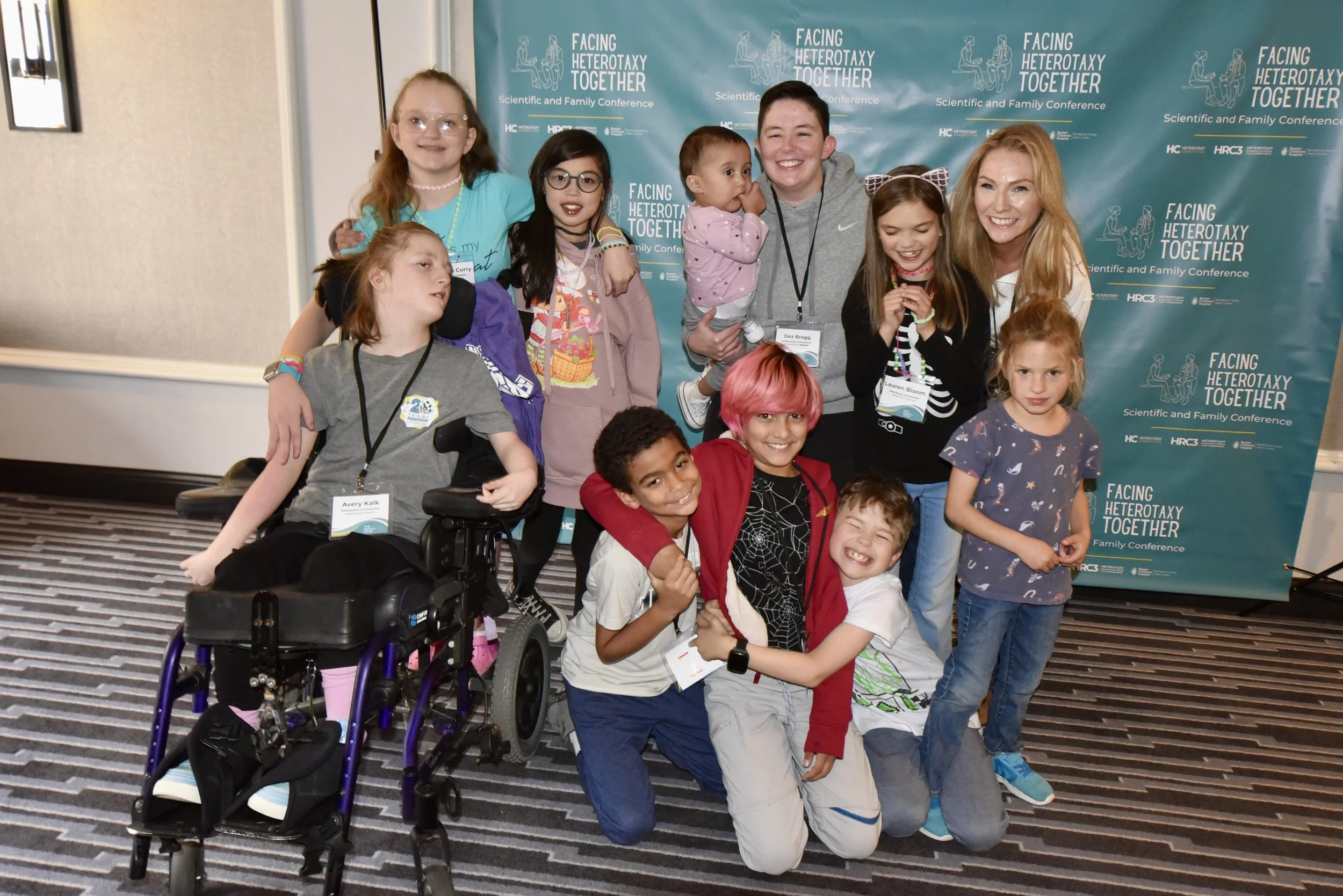 A diverse group of children of various ethnicities and disabilities and several adults stand and sit smiling in front of a teal step and repeat banner