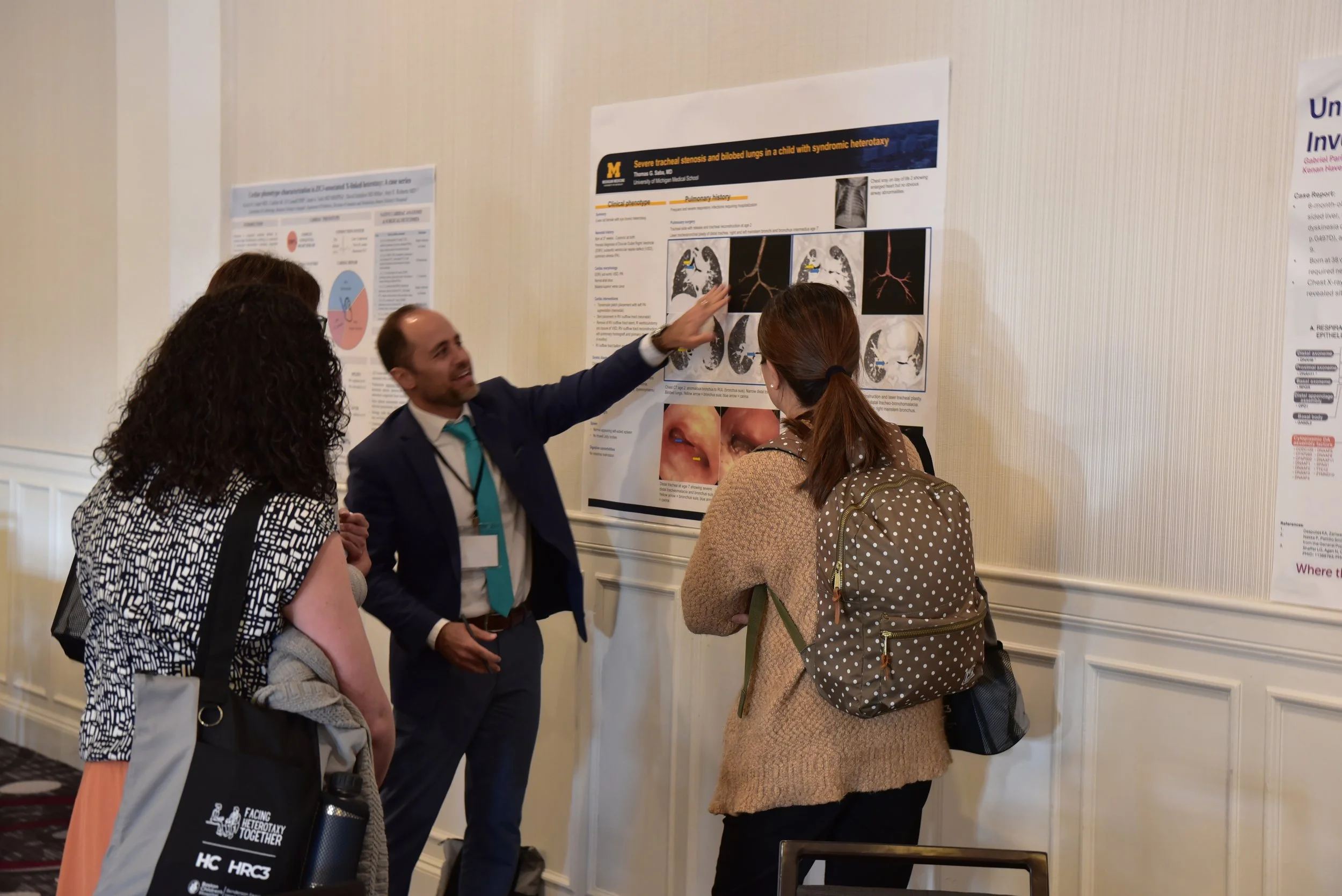 a man in a suit points to a poster containing medical research while two women look on