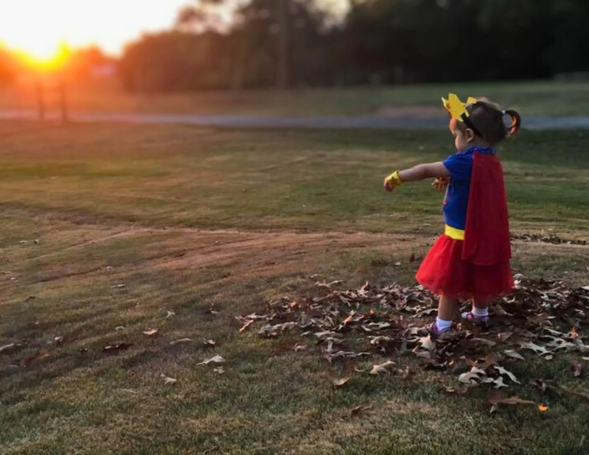 A toddler girl in a superhero costume stands on the right side of the photo, arms outstretched towards the glowing sunset on the left.