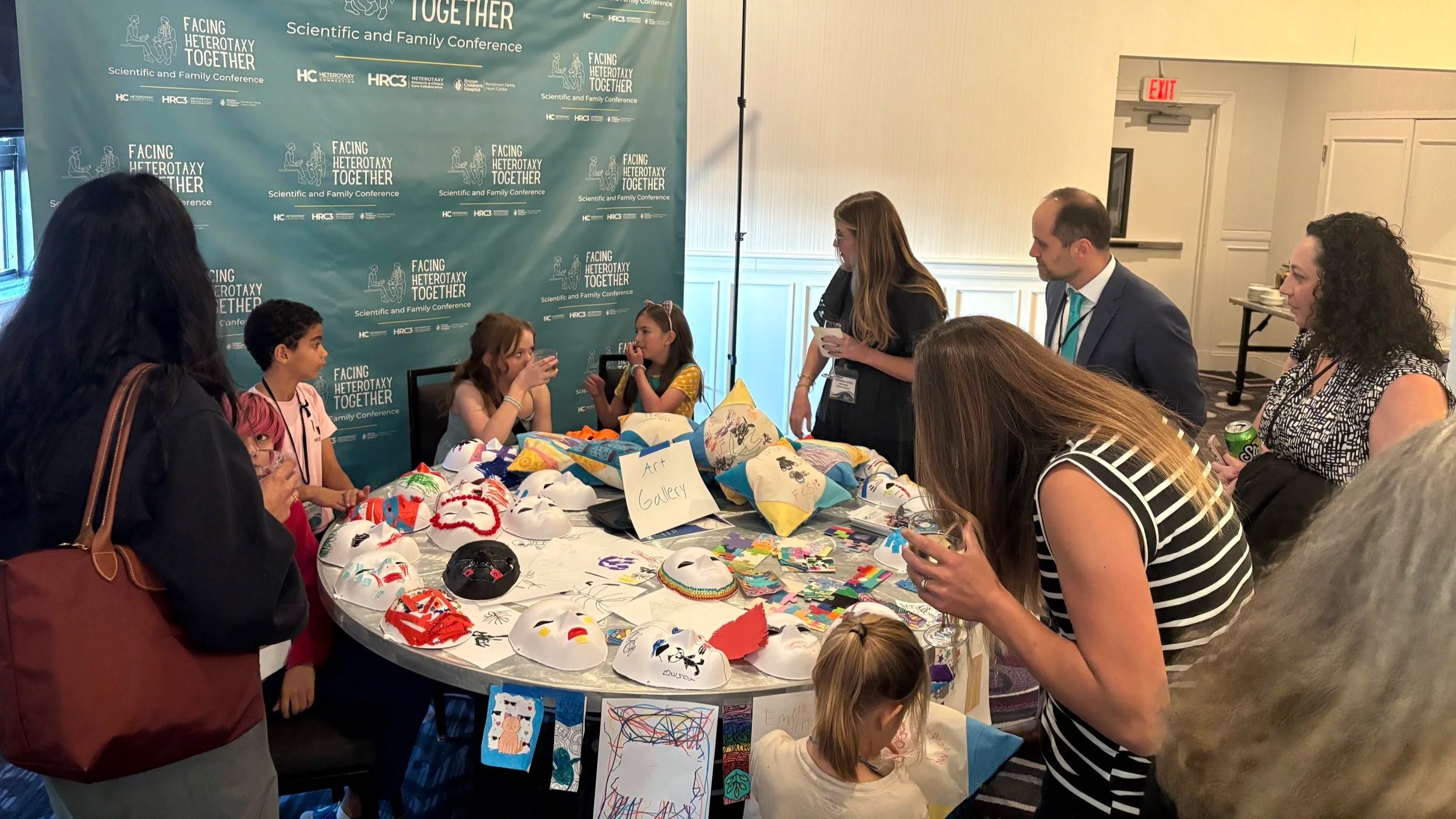 At round table covered with arts and crafts, several children sit ready to answer questions while adults inspect the artwork and engage the children.
