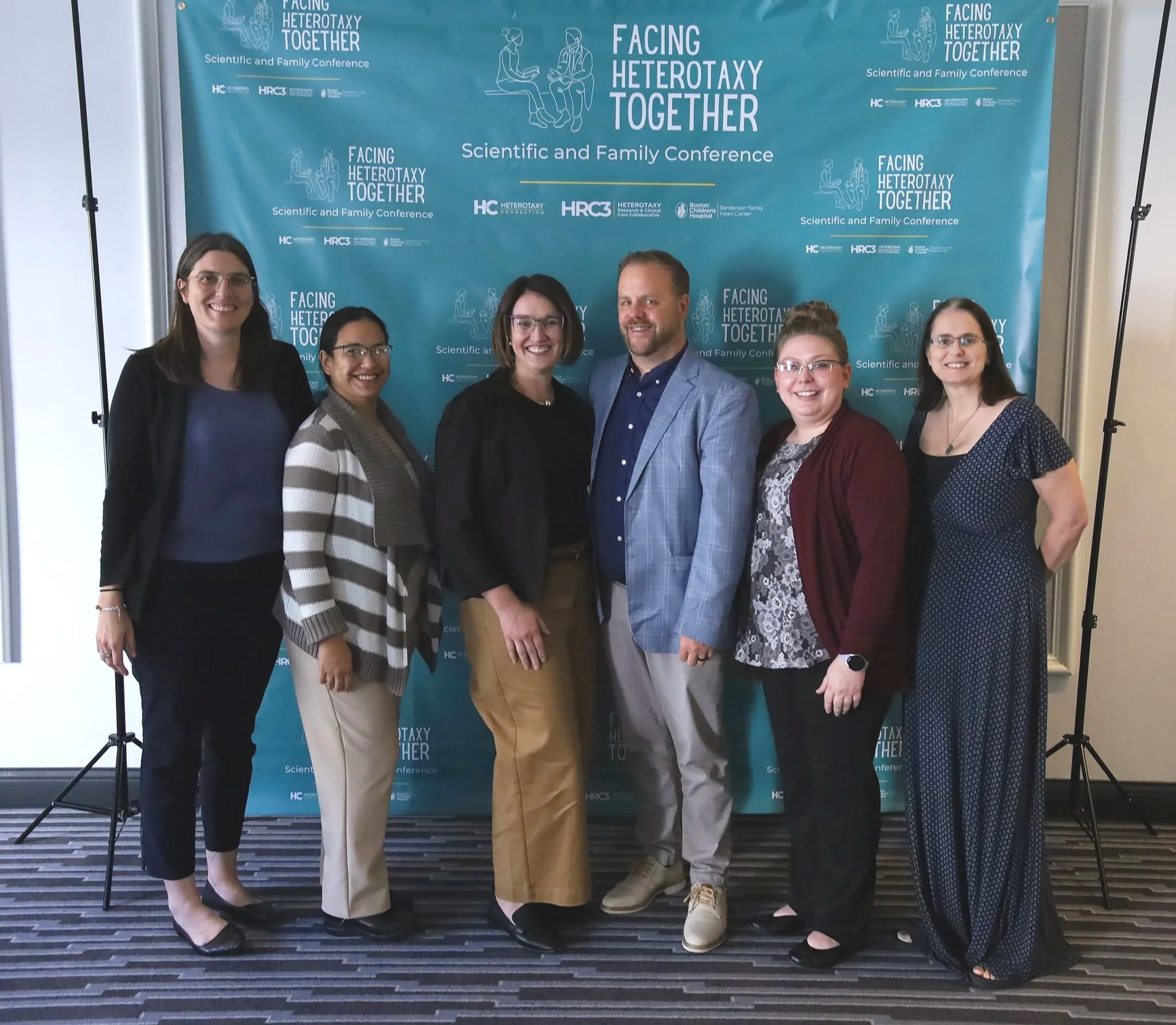 6 people, 5 women and one man, stand smiling in front of a teal step and repeat banner with the Facing Heterotaxy Together logo.