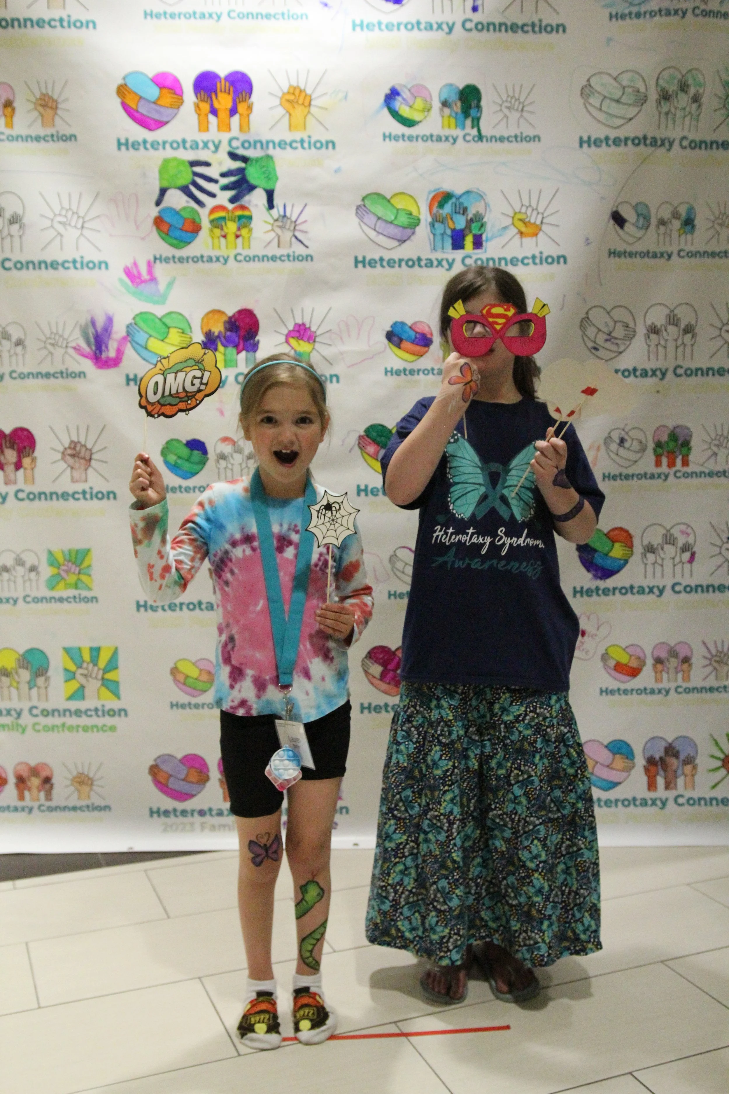 two young girls stand in front of a brightly colored step and repeat banner. They are smiling and holding superhero photobooth props