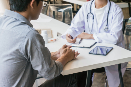 A man in a lab coast with a stethoscope sits across the table from another man in a button down shirt. The man in the lab coat is taking notes.