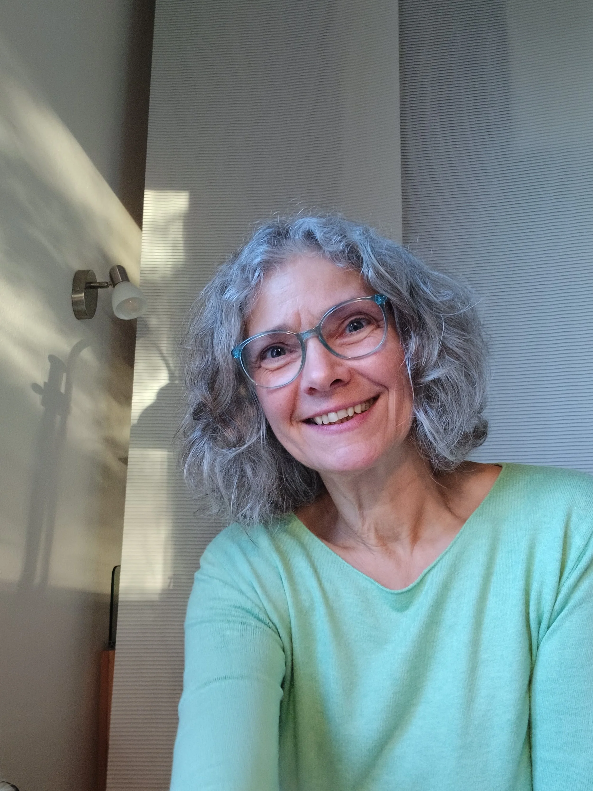 Ein lächelnder älterer Mensch mit lockigem grauem Haar und Brille, getragen in einem hellgrünen Oberteil, bei Tageslicht in einem Raum mit Lampe und weißen Vorhängen.