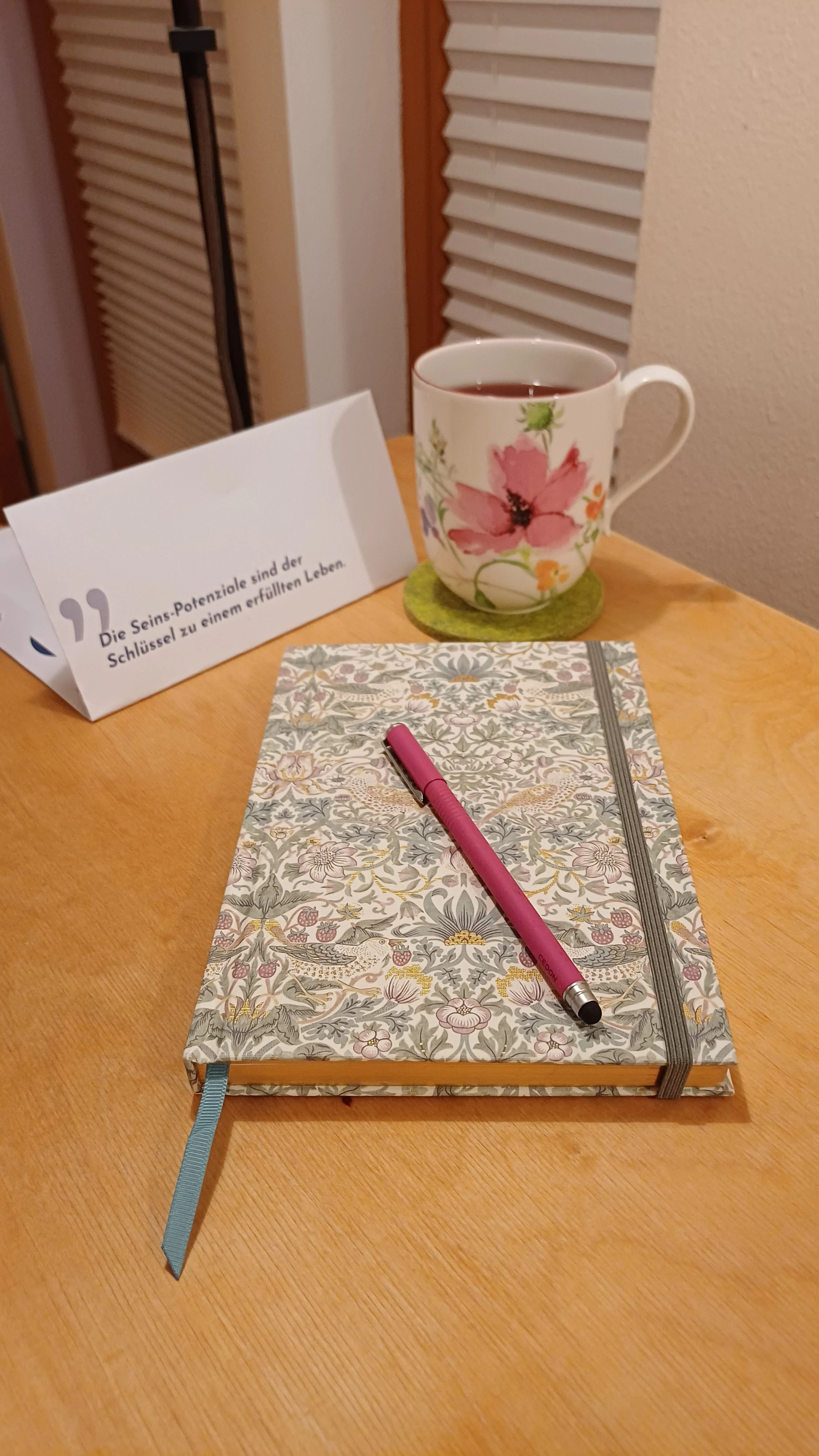 Tagebuch mit Blumenmuster, rosa Kugelschreiber, Tasse mit Blumenmuster und Tee, Notizkarte mit Zitat auf einem Holzschreibtisch.