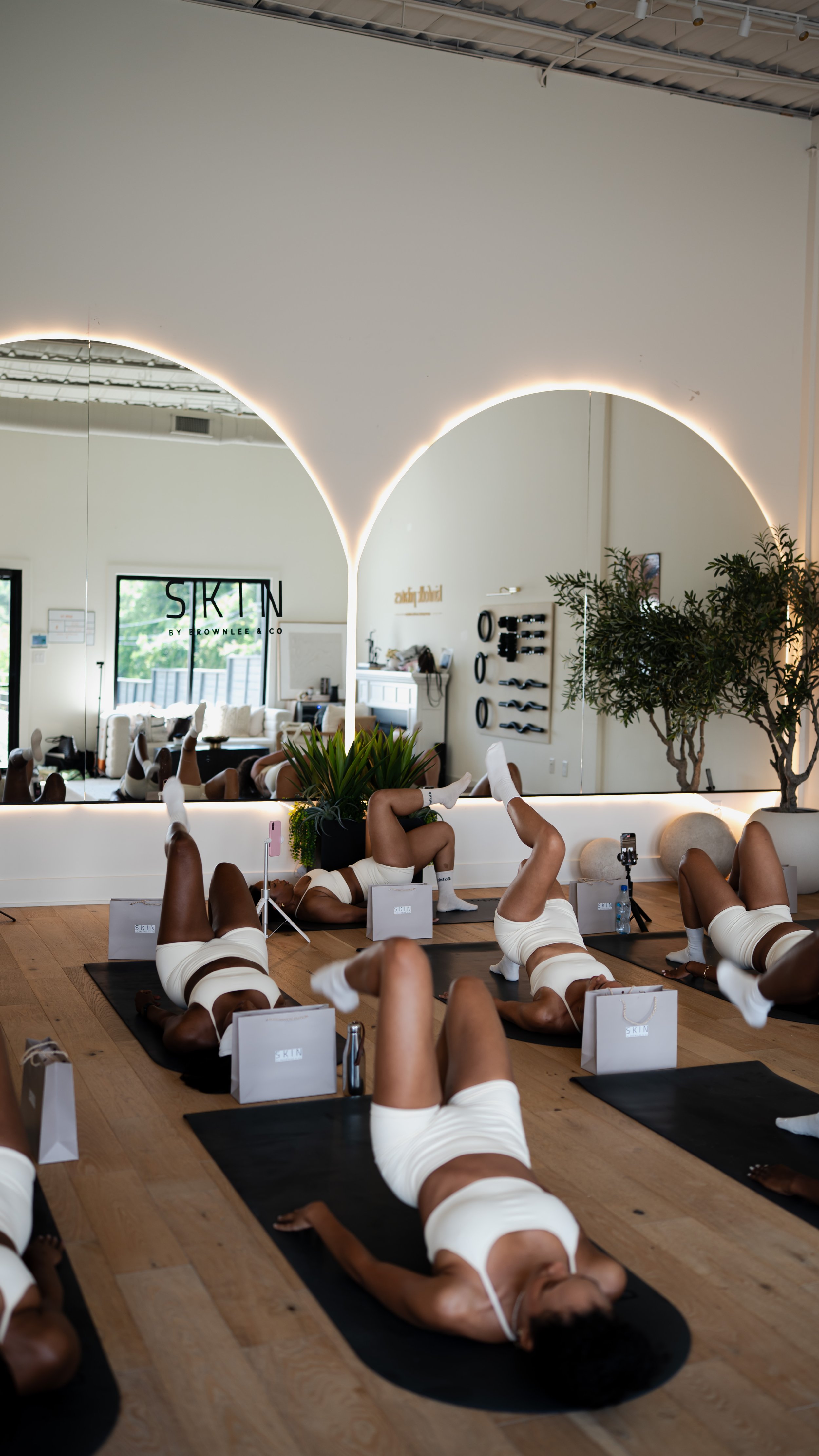 A pregnant woman practicing yoga indoors, stretching with one arm raised and holding her foot behind her, wearing a white sports bra and white leggings.