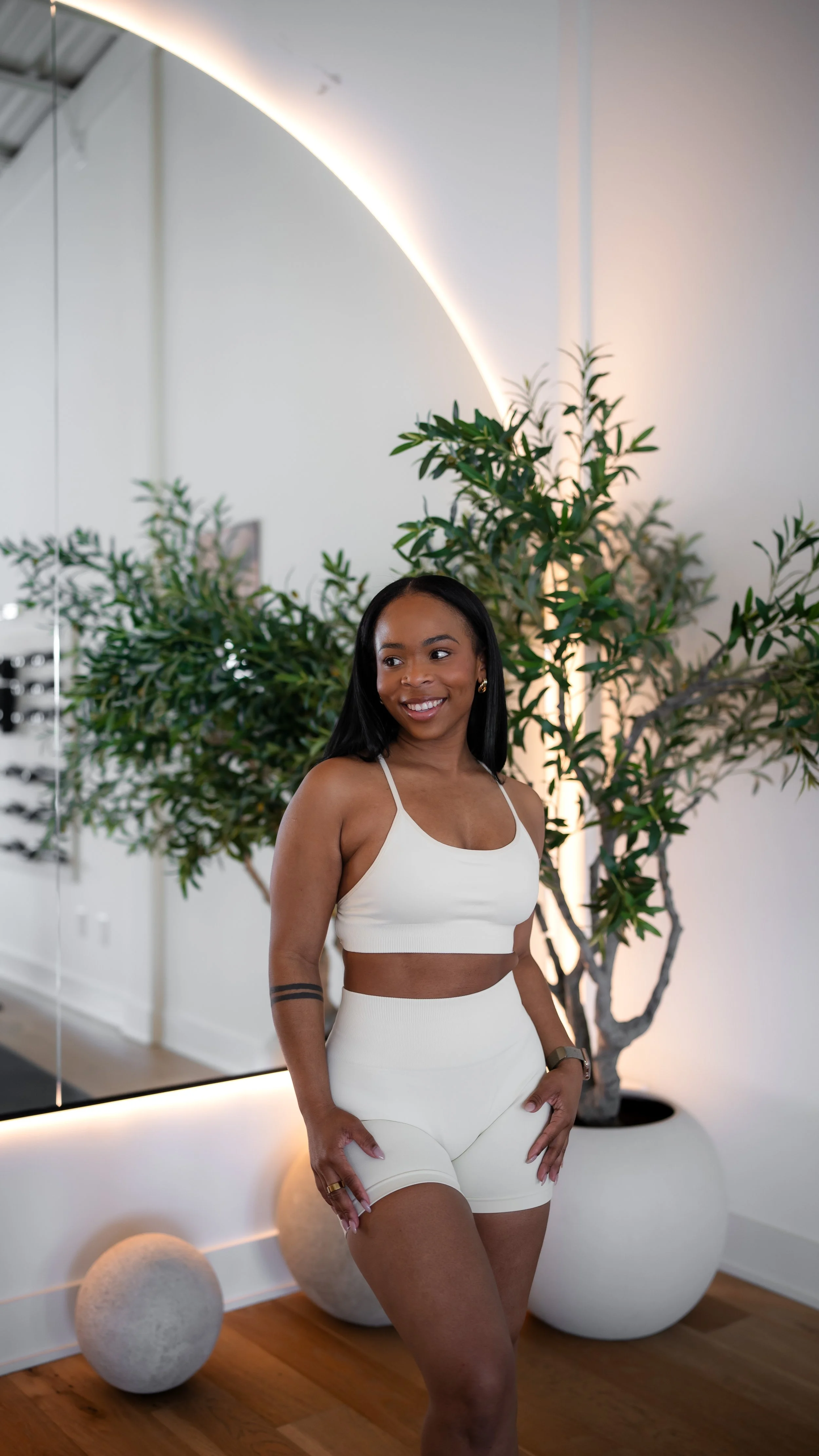 A woman in a white athletic crop top and high-waisted shorts standing indoors near a large potted plant and a mirror, smiling.