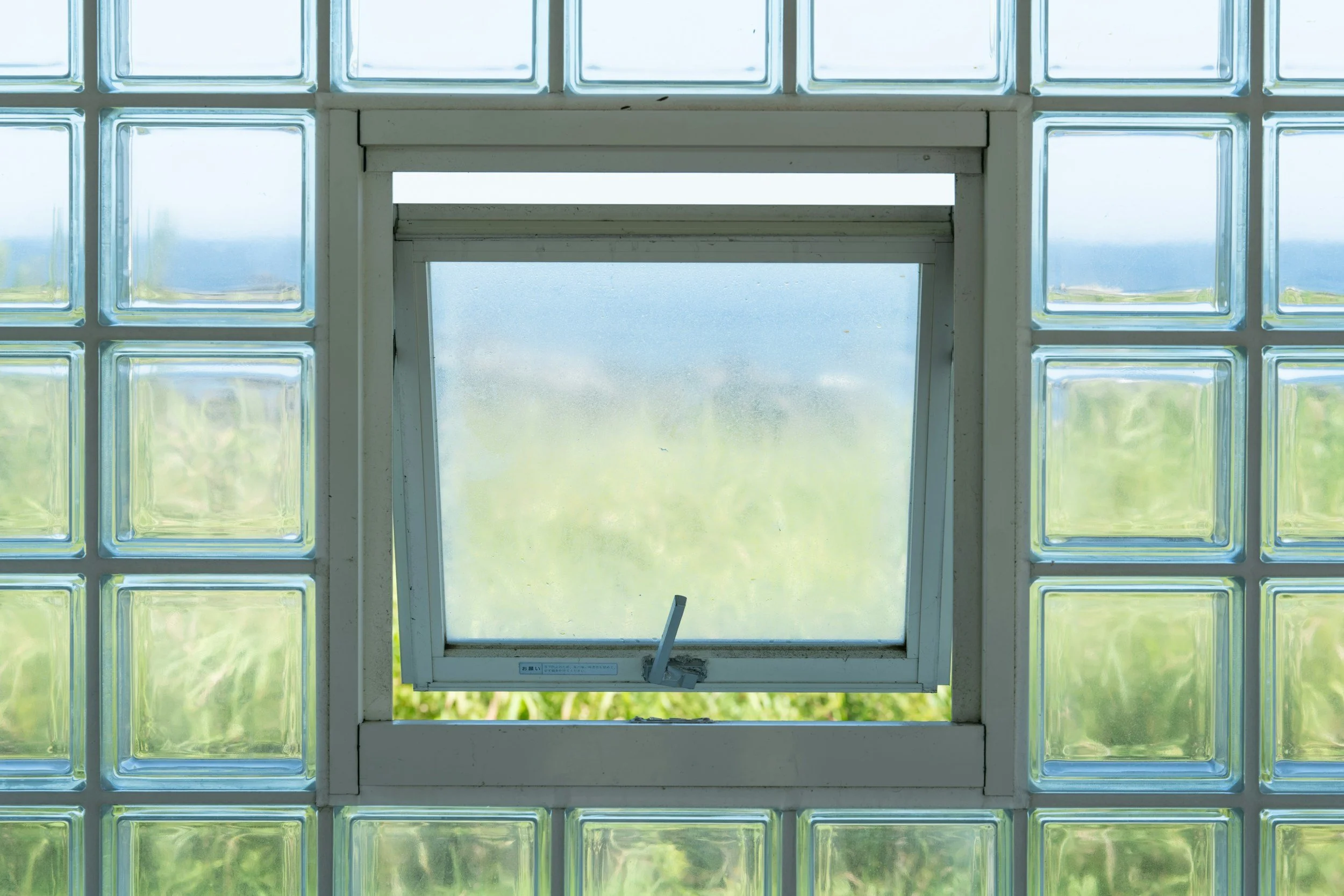Open window with frosted glass blocks surrounding it, overlooking a green landscape and blue sky.