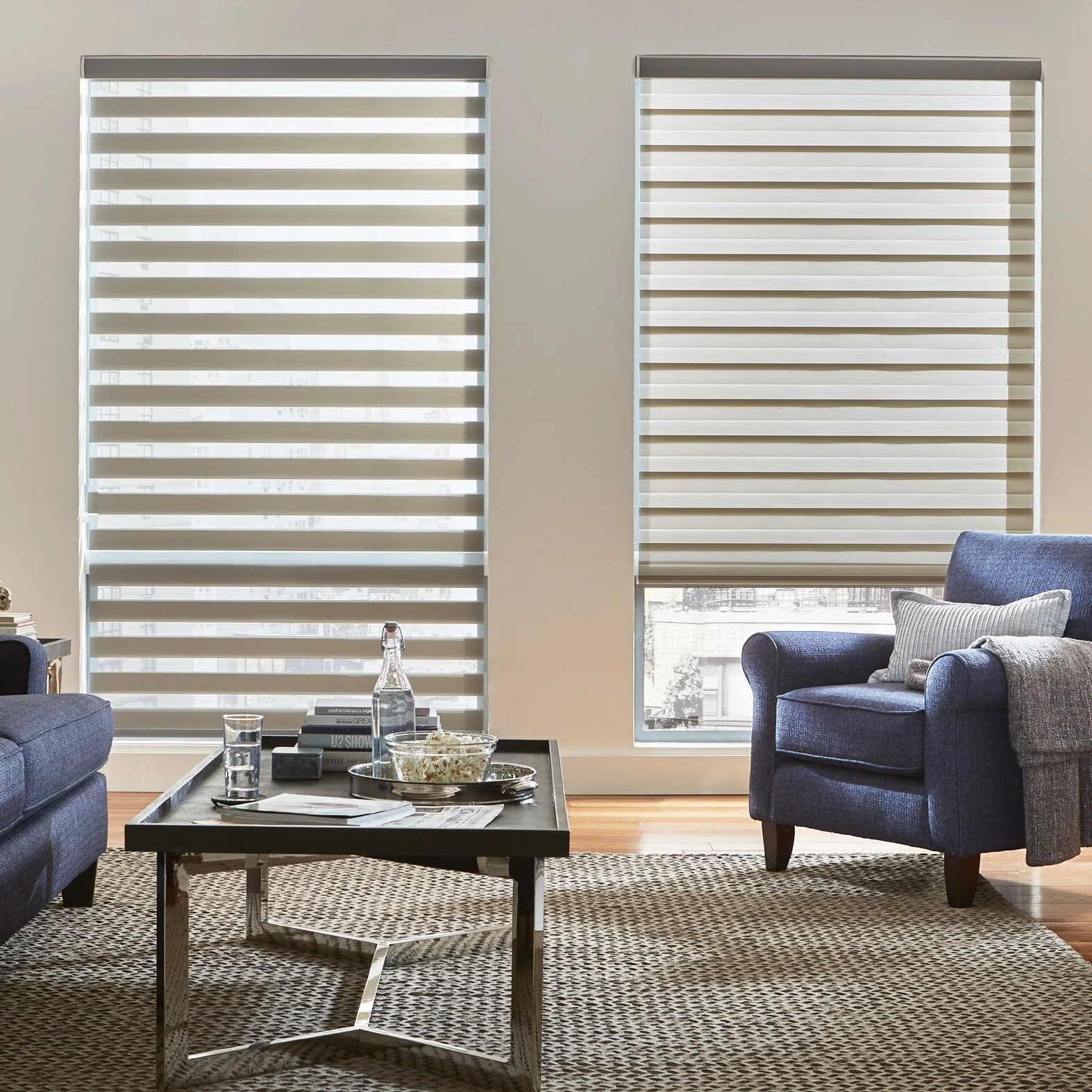 Living room with two large windows covered by horizontal blinds, blue armchair with pillows, patterned rug, coffee table with snacks and drinks.