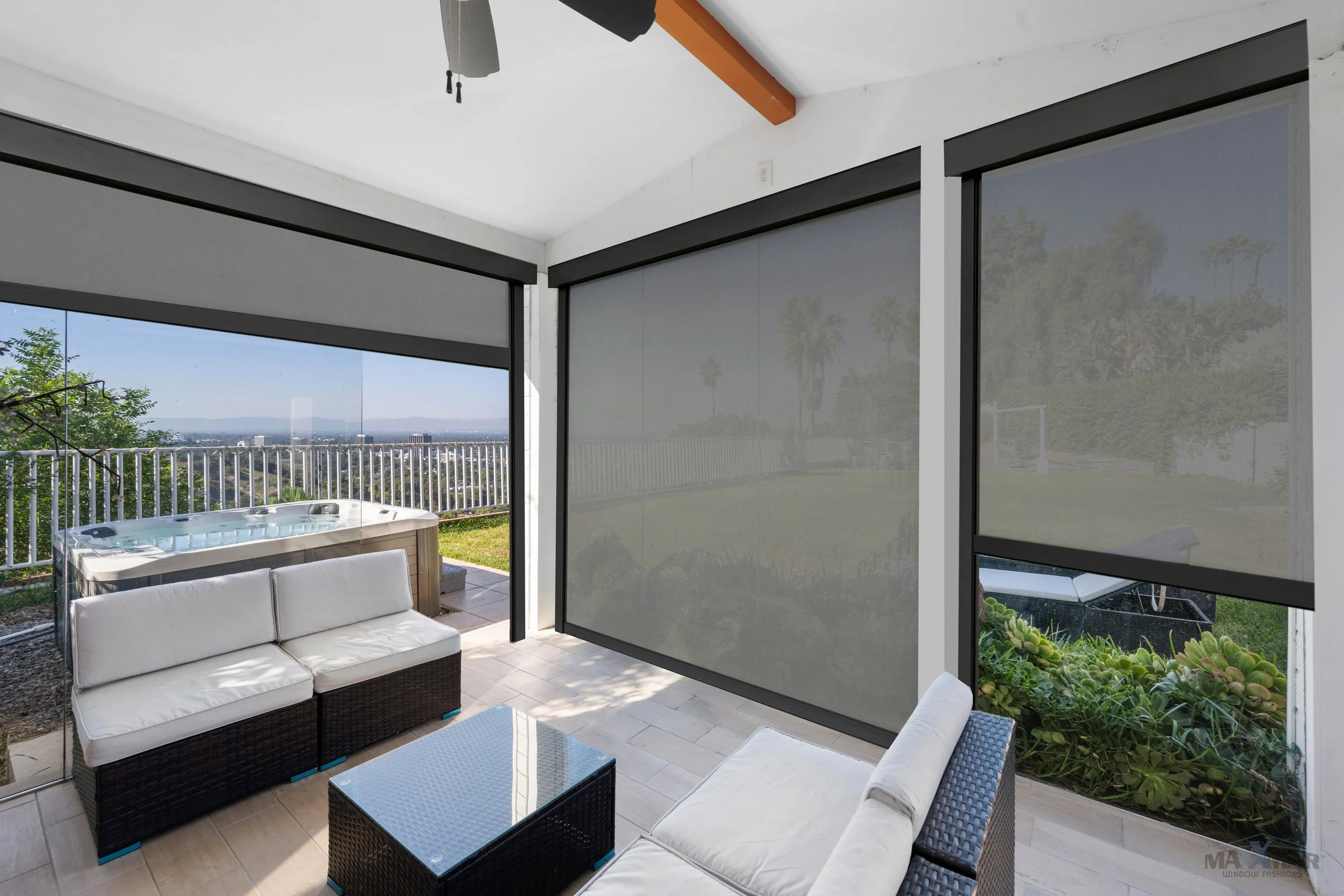 A screened-in patio with outdoor furniture, including a white cushioned sofa, two white cushioned chairs, and a glass-top table, overlooking a backyard with a hot tub and a view of distant cityscape and palm trees.