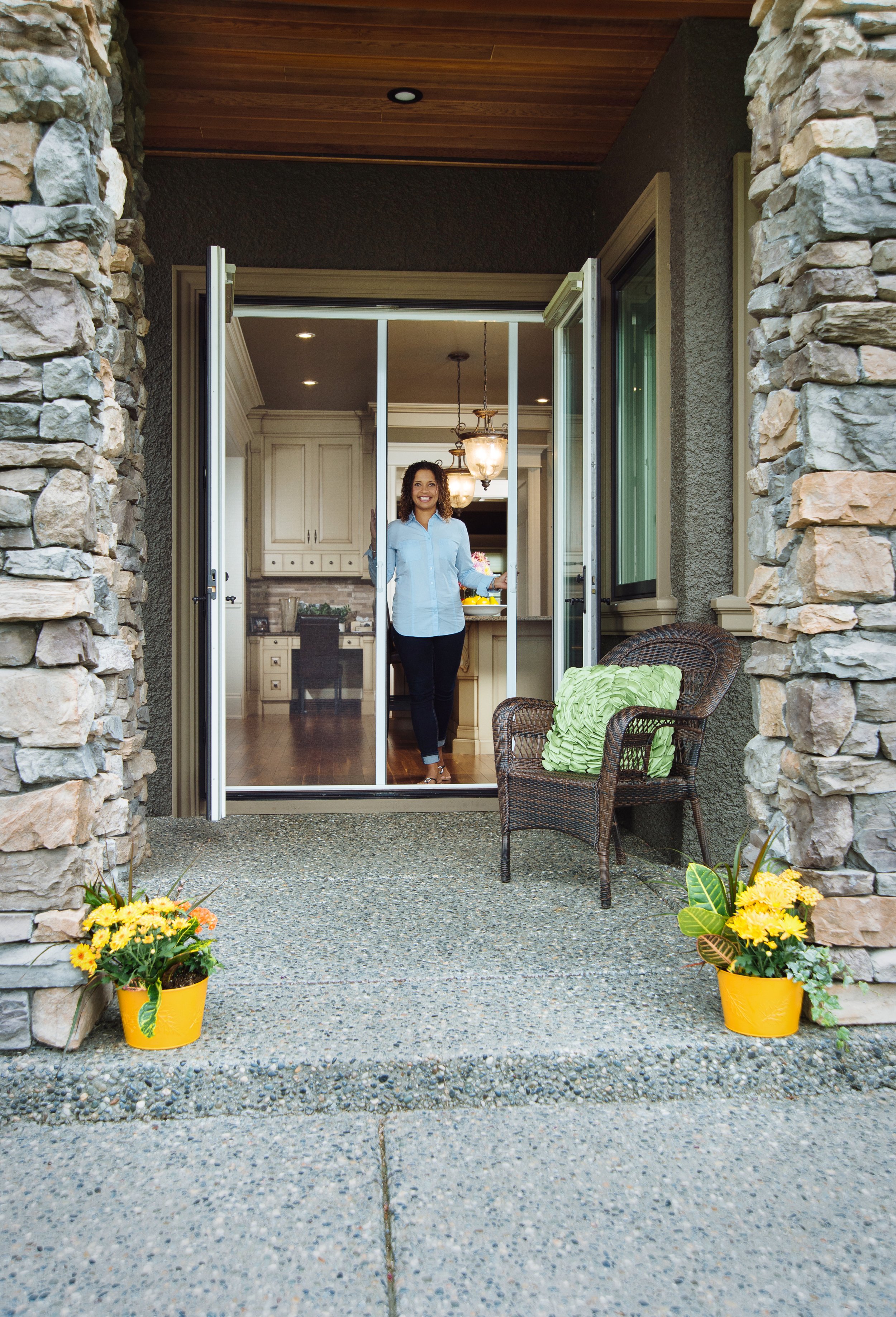 A woman standing inside a house near a sliding glass door, smiling, with a stone porch and potted flowers outside.