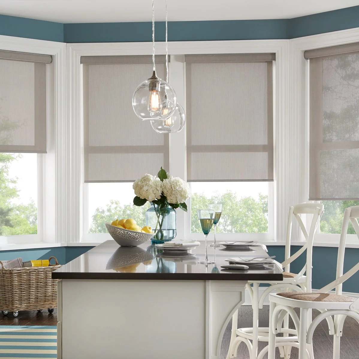 A dining area with a kitchen island, white chairs, a blue wall, and four windows with gray blinds. There is a vase with white flowers and a bowl of lemons on the island, along with glasses of white wine and plates.