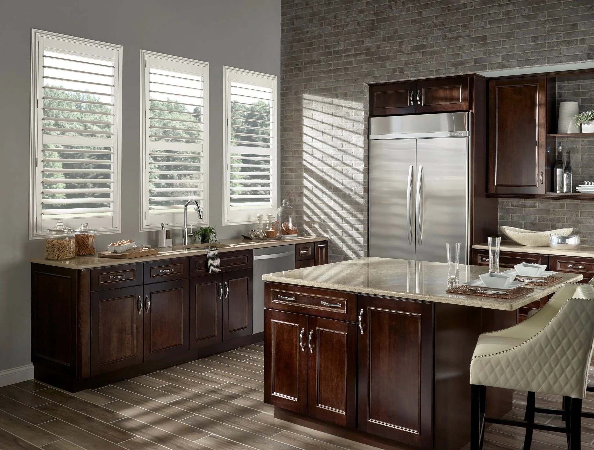 Modern kitchen with dark wood cabinets, a beige marble island, stainless steel refrigerator, gray brick wall, wood floor, and three white window shutters.