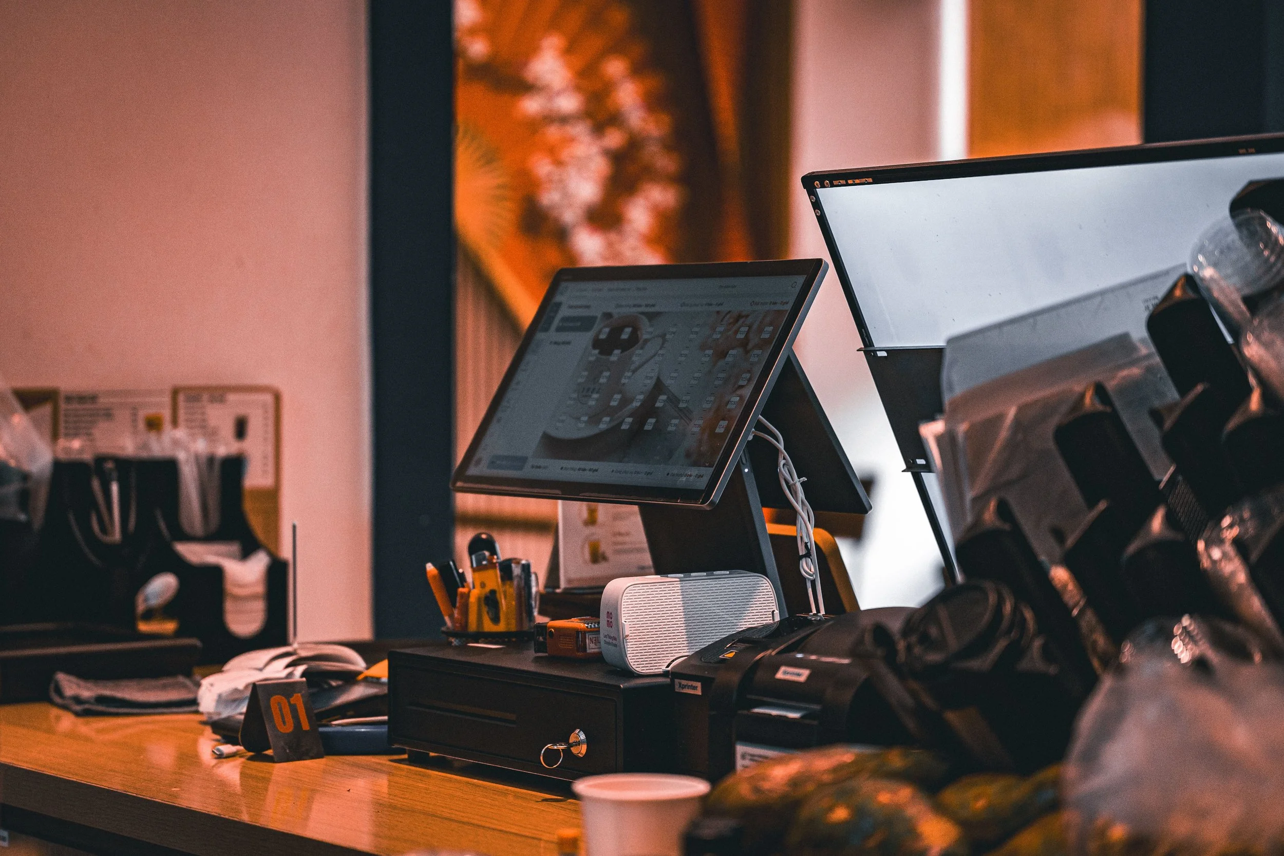 A cluttered office workspace with multiple computer monitors, a keyboard, various office supplies, and papers scattered on a wooden desk. The background features a blurred wall with a framed picture or poster.