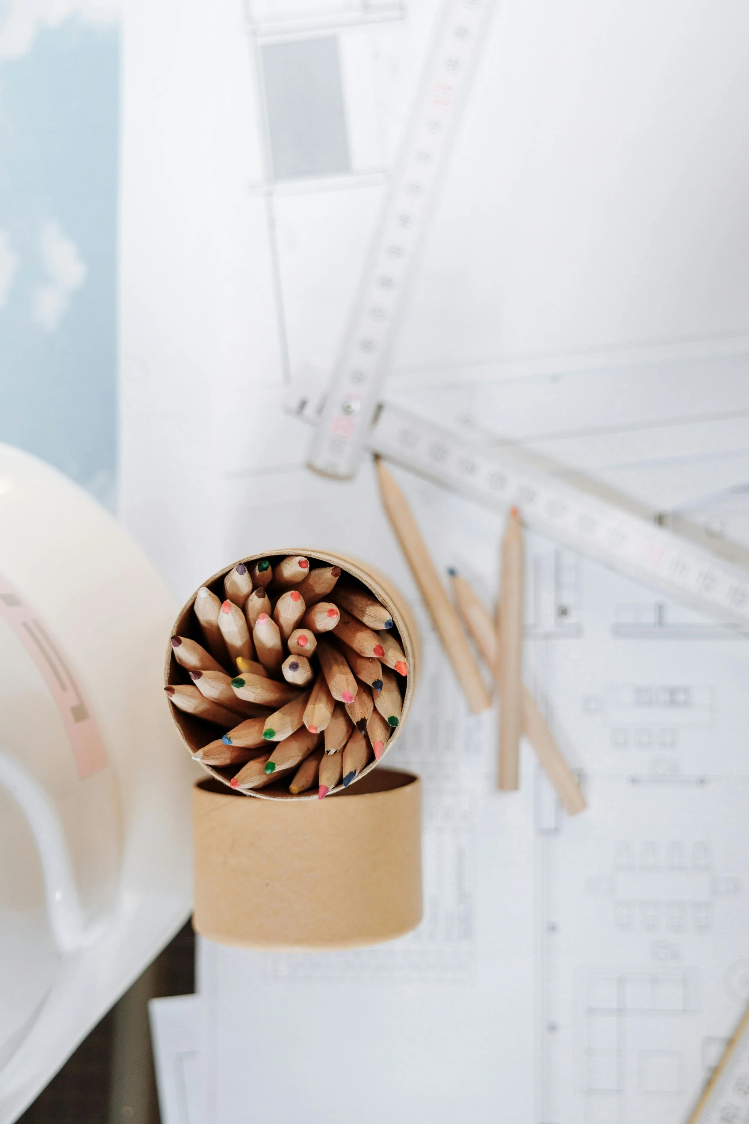 A top-down view of a pencil holder filled with colored pencils, placed on a table with architectural blueprints, rulers, and wooden pencils.