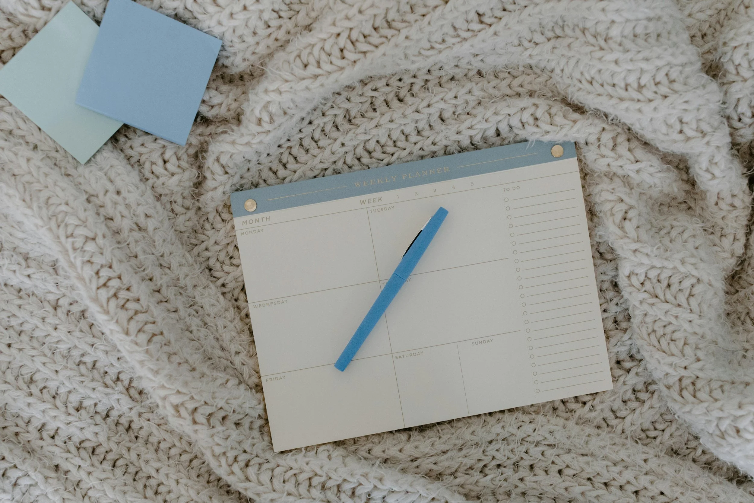 A weekly planner, a blue pen, and two sticky notes on a cream knitted blanket.