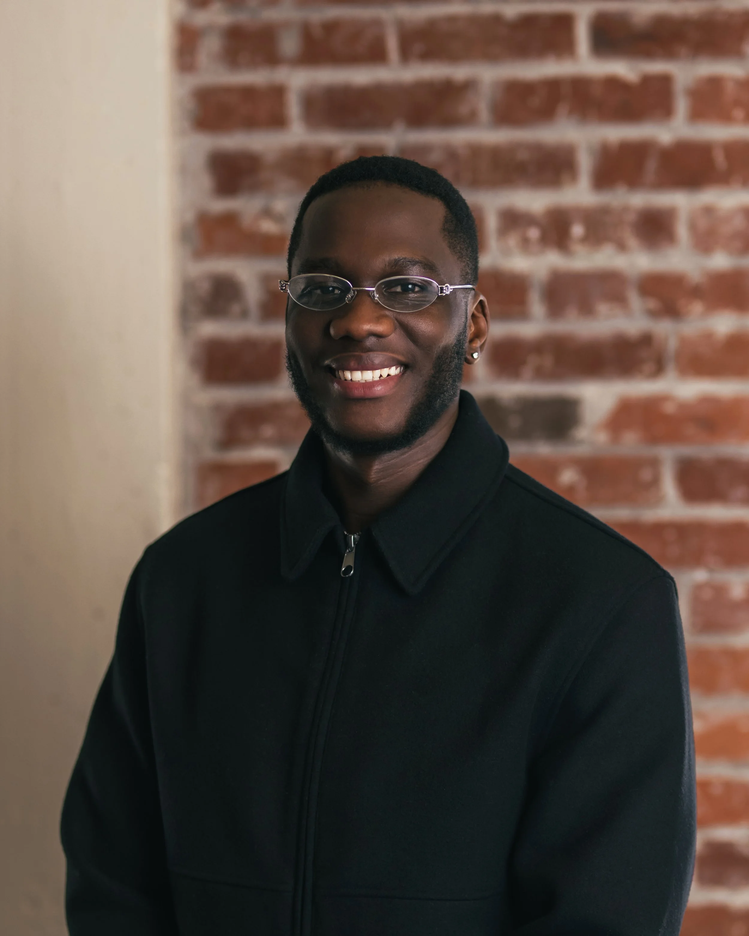 professional portrait of a man wearing glasses, smiling, in a black zip-up jacket, against a brick wall background