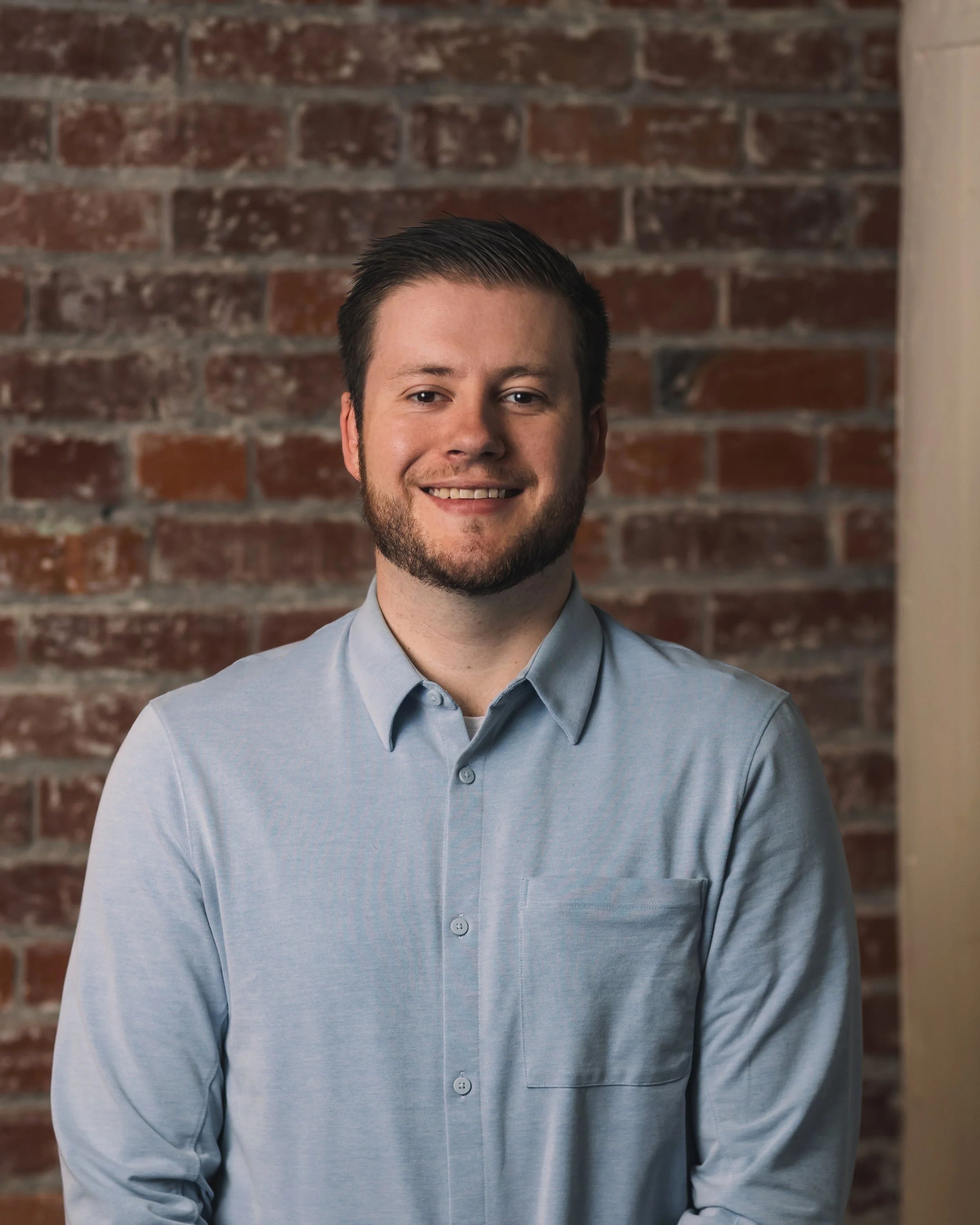 professional portrait of a man with short brown hair and a beard, smiling, wearing a light blue button-up shirt, against a brick wall background