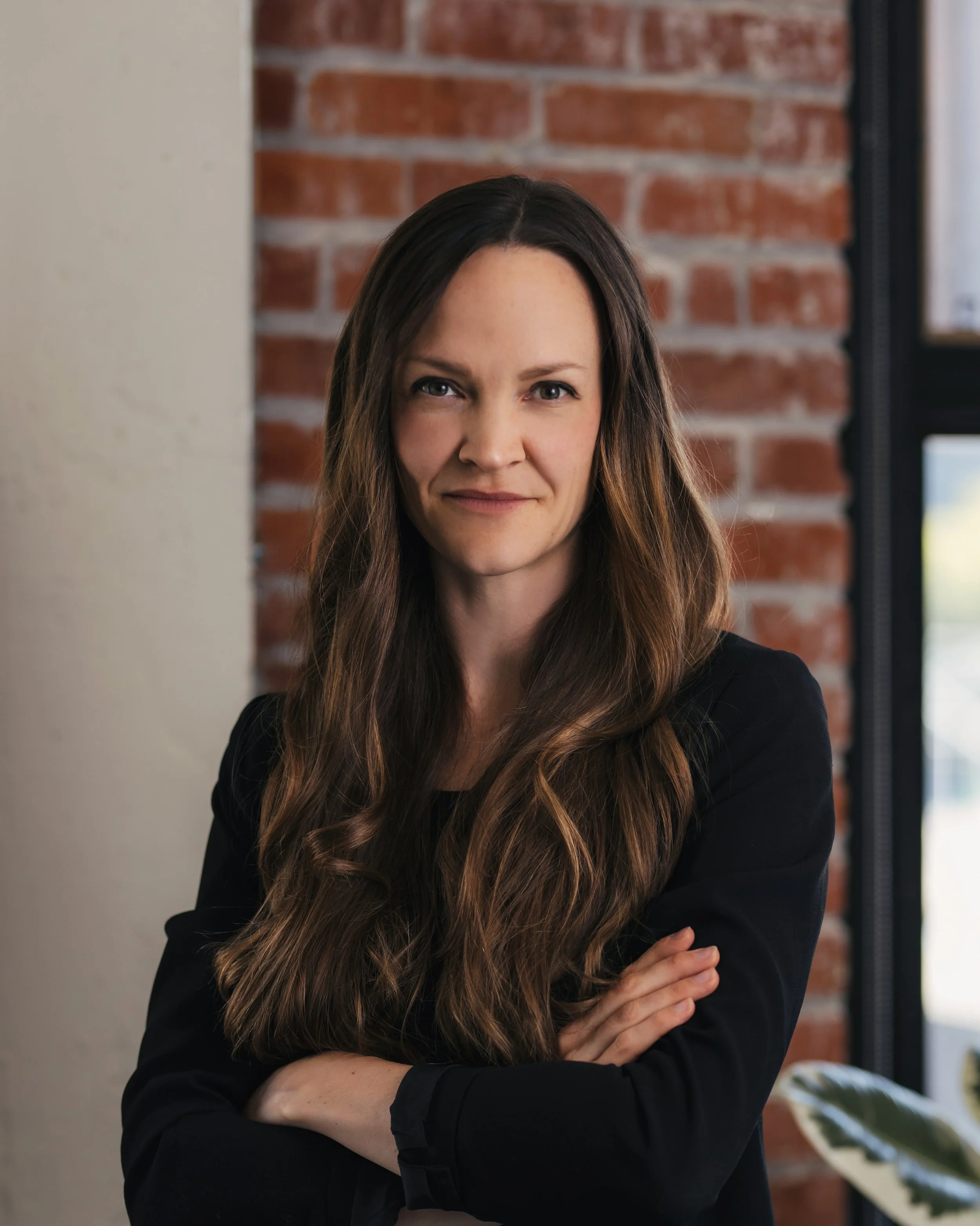 professional portrait of a woman with long brown hair, arms crossed, smiling confidently against a brick wall background