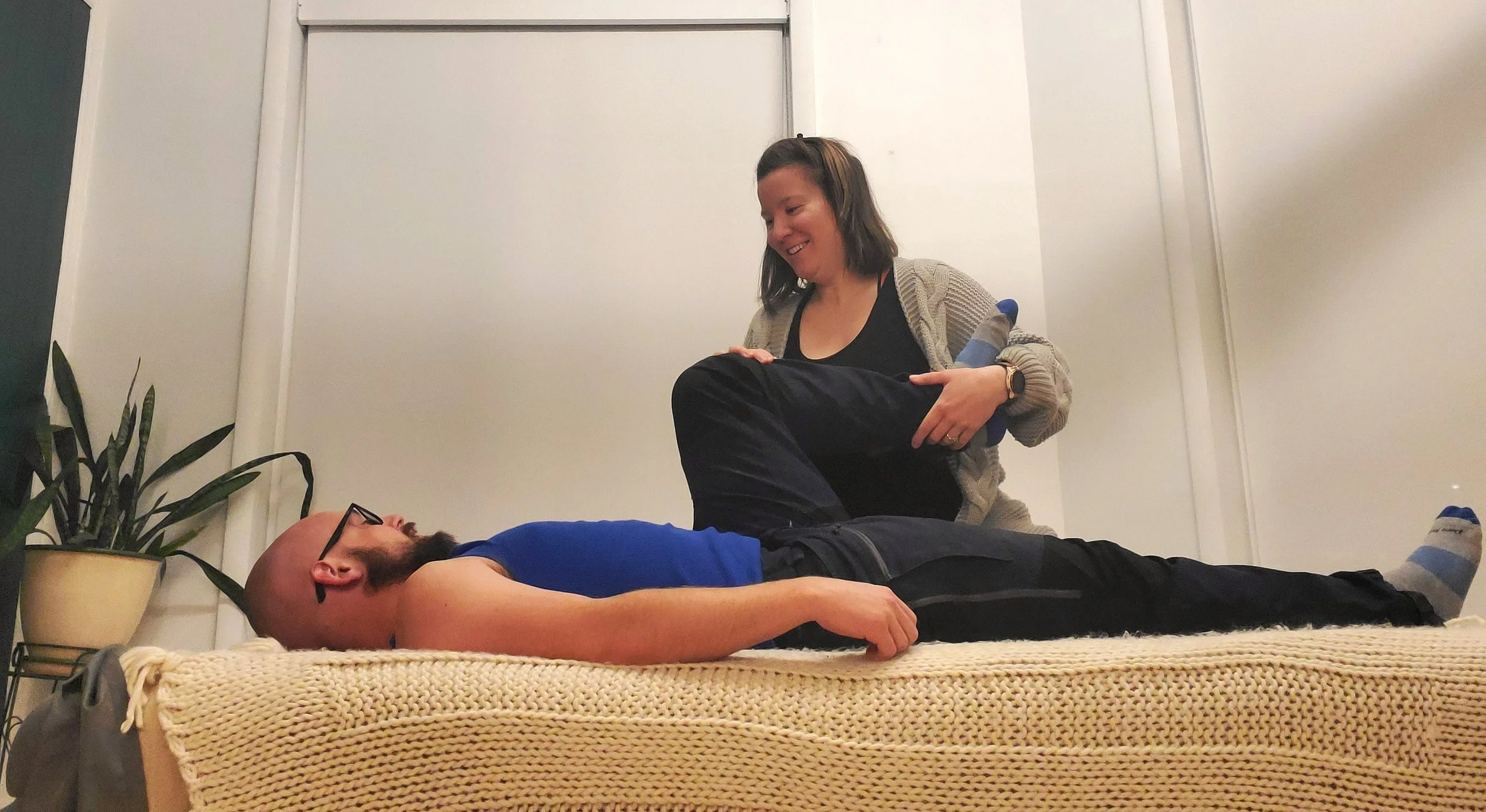 A woman sitting on a bed with a man lying down on the mattress. The woman is smiling and looking at the man, who is resting with his eyes closed.